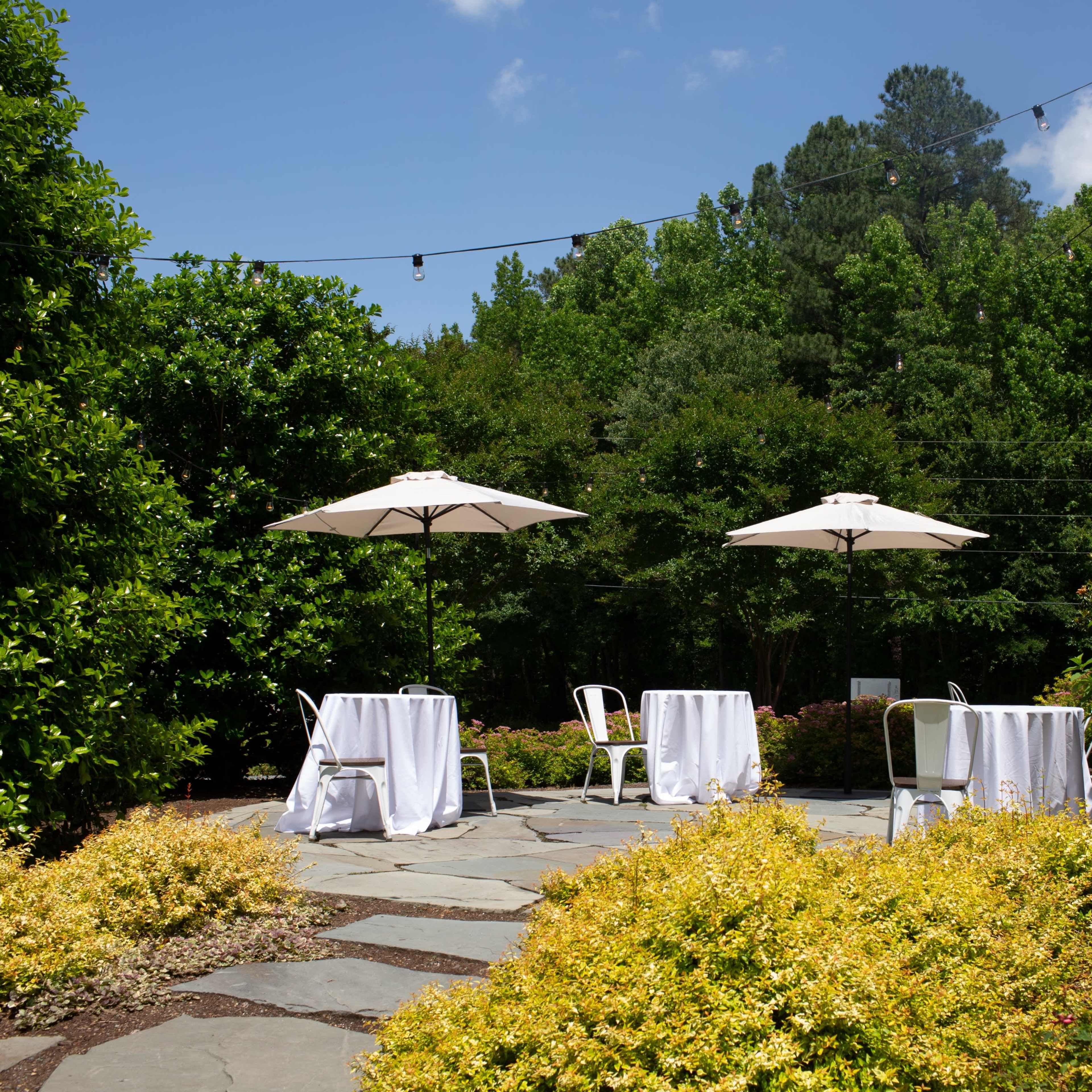 The scene features two tables covered with white tablecloths under umbrellas, set on a stone pathway surrounded by lush greenery and vibrant yellow plants.
