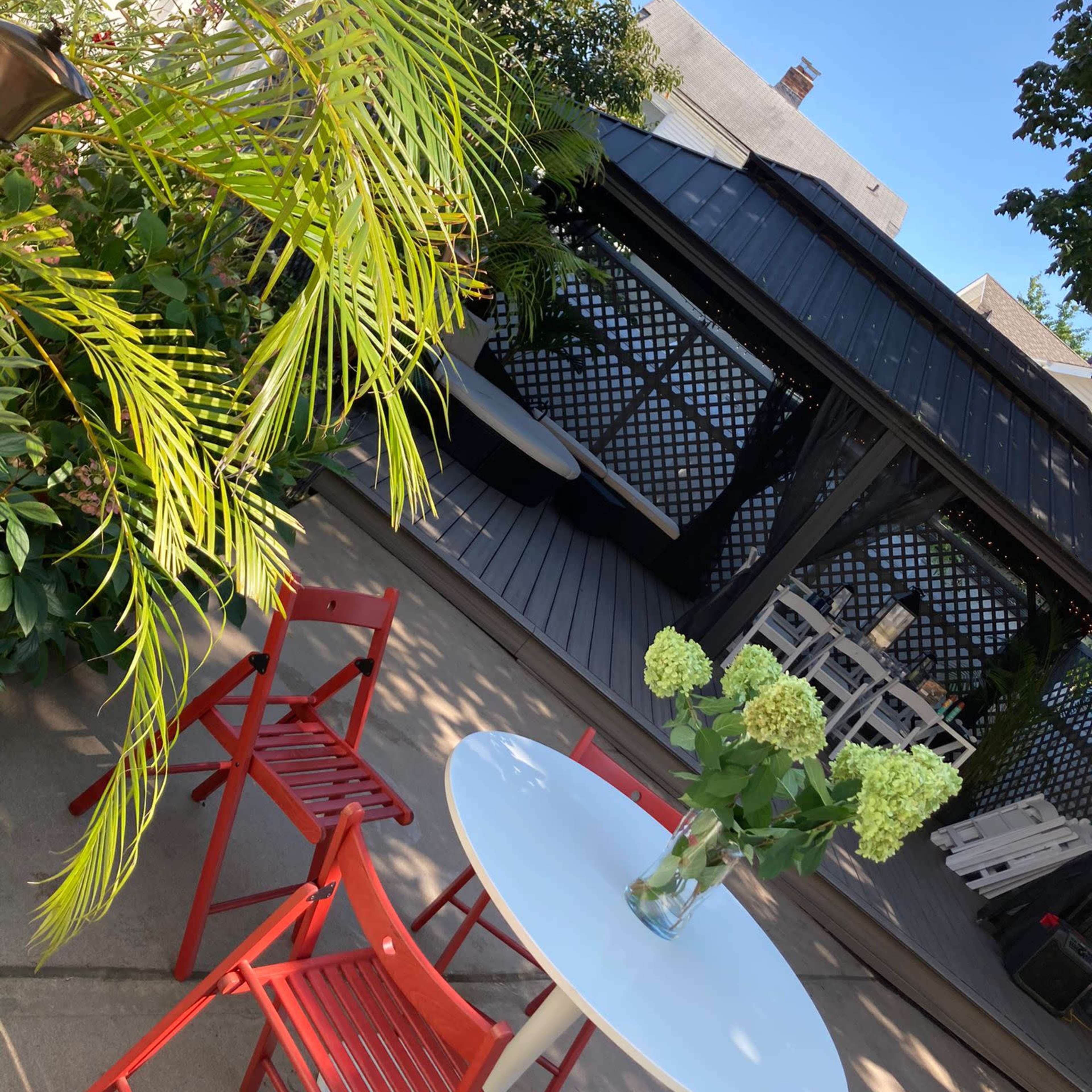 A patio area with a round white table and red chairs, surrounded by greenery and a shaded seating space in the background.