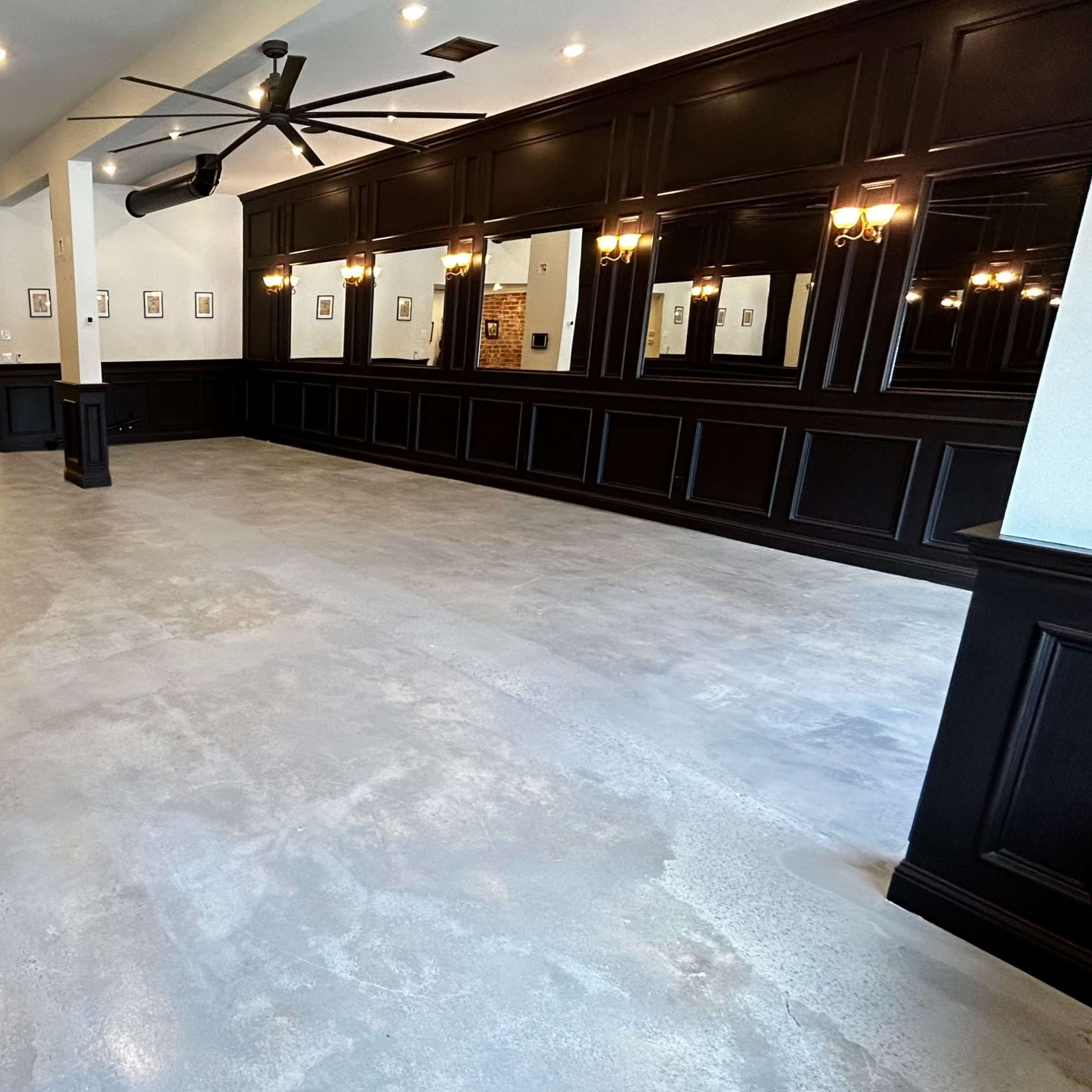 A spacious interior room with polished concrete flooring and dark wood paneling, featuring large mirrors and ceiling fans.