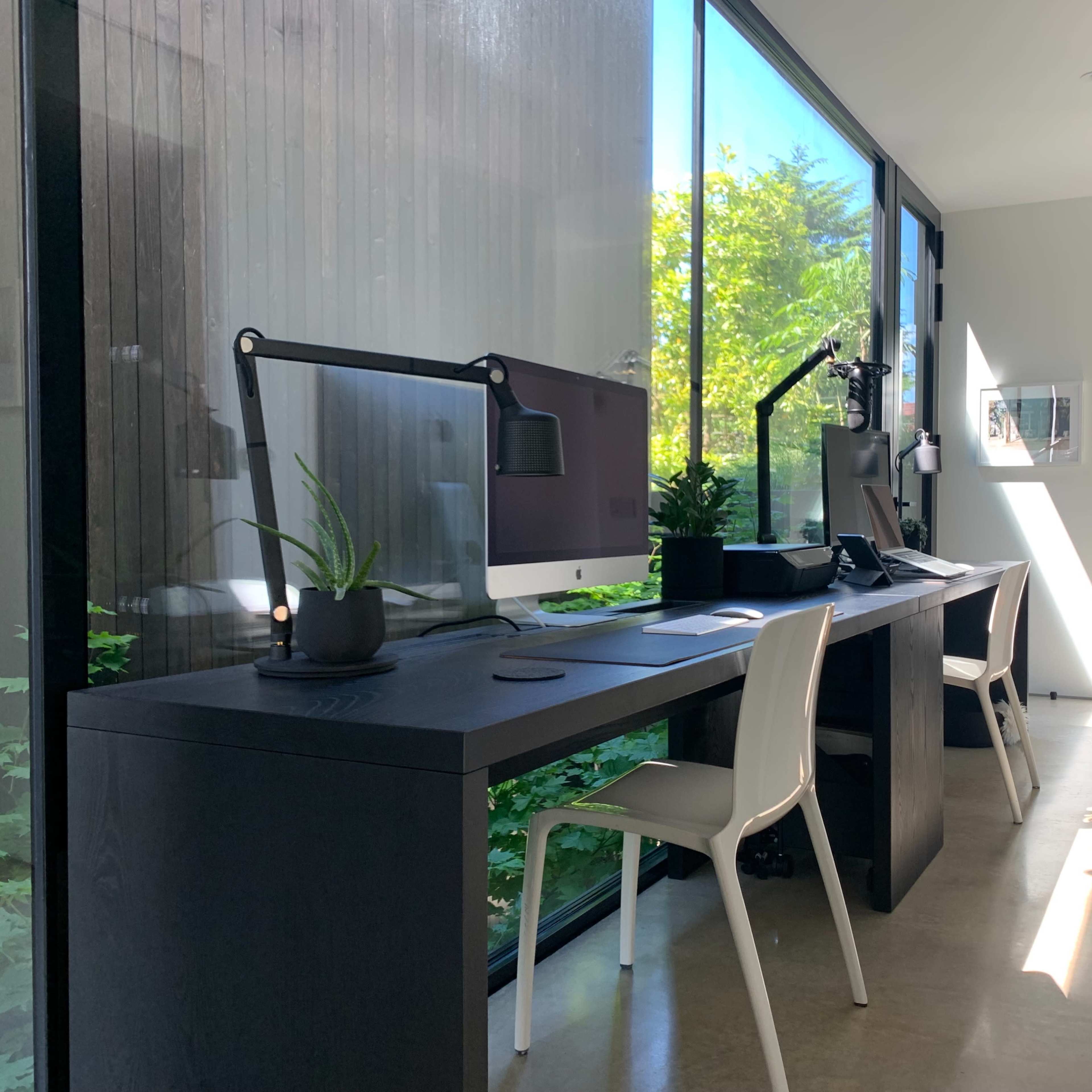 A modern home office featuring two desks with computers, a potted plant, and large windows allowing natural light to enter.
