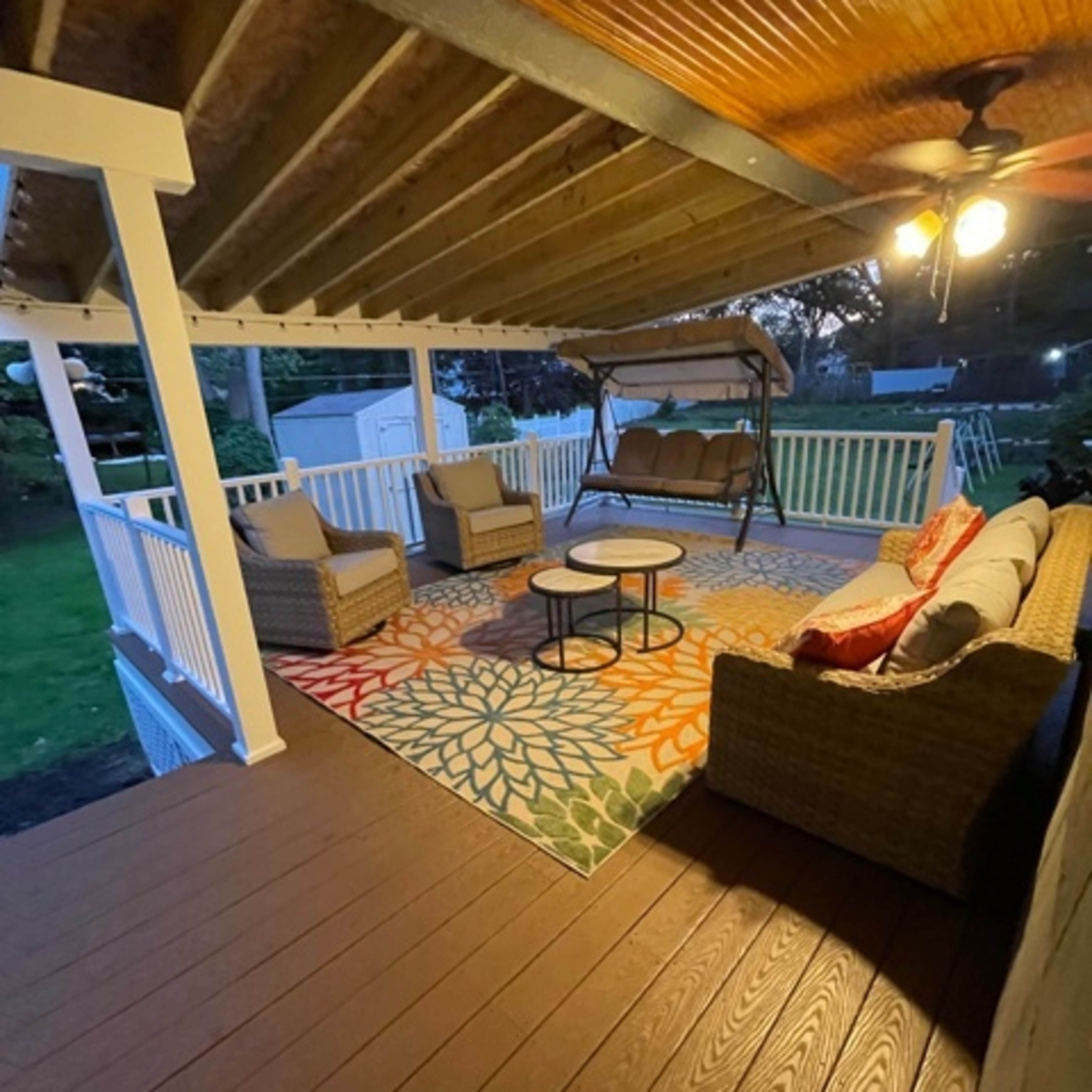 A covered patio area furnished with a swing, two chairs, a couch, and a round coffee table on a colorful patterned rug.