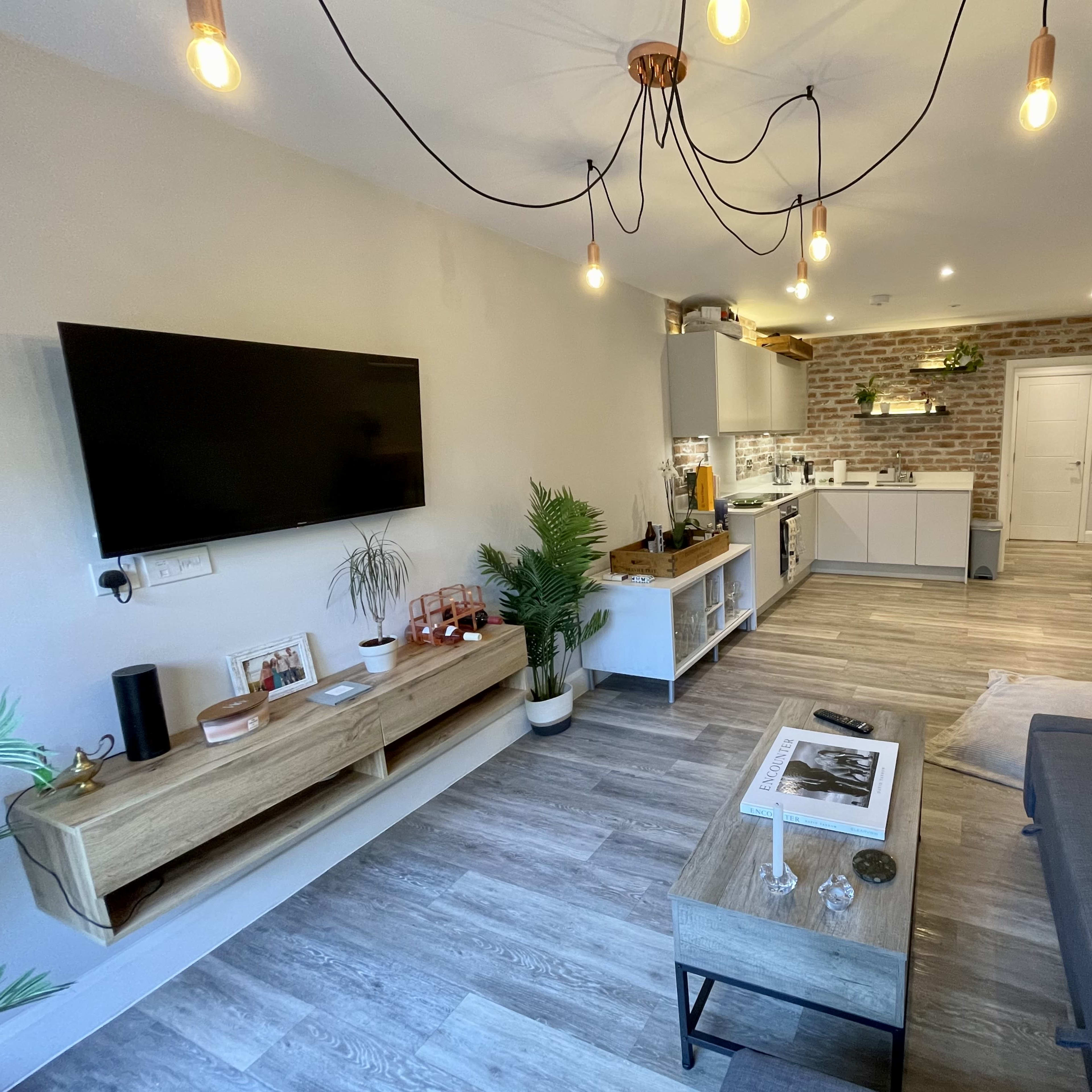A modern living room with a gray sofa, a wooden coffee table, a TV mounted on the wall, and a kitchen area with brick accents visible in the background.