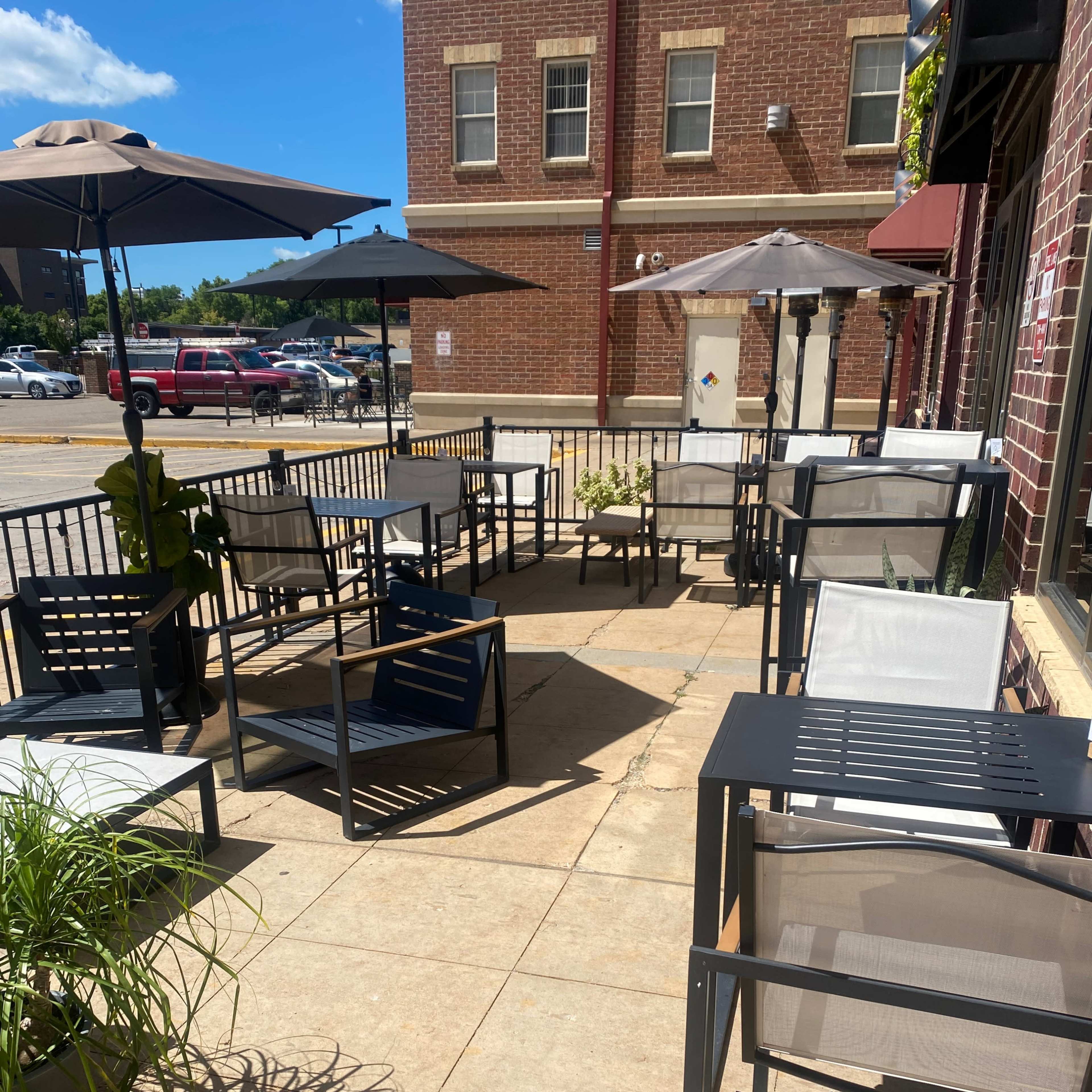 A patio area with several empty, modern black and gray chairs and tables, surrounded by umbrellas and potted plants.