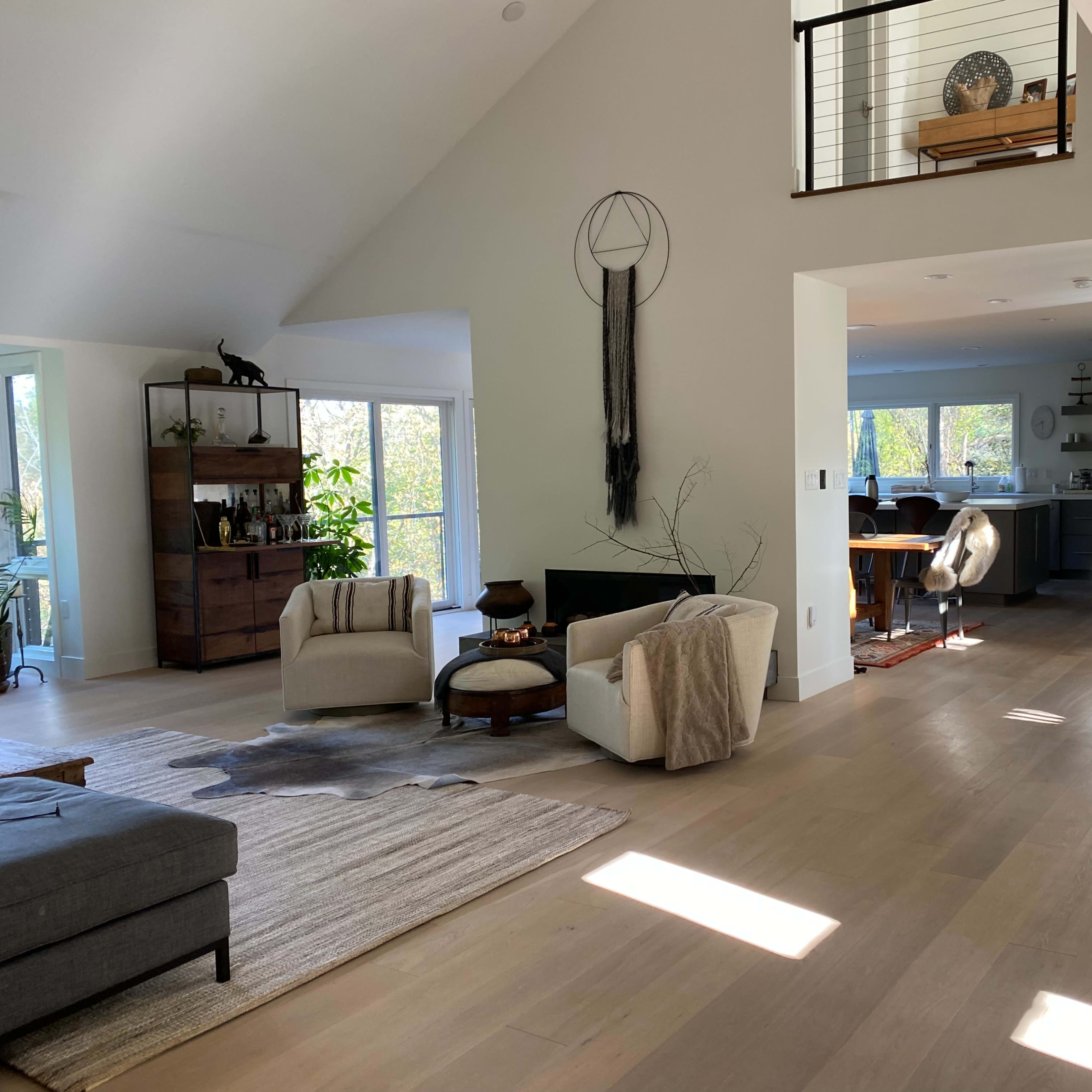The interior of a modern home features an open layout with a living area, dining space, and kitchen visible, adorned with plants and natural light from large windows.