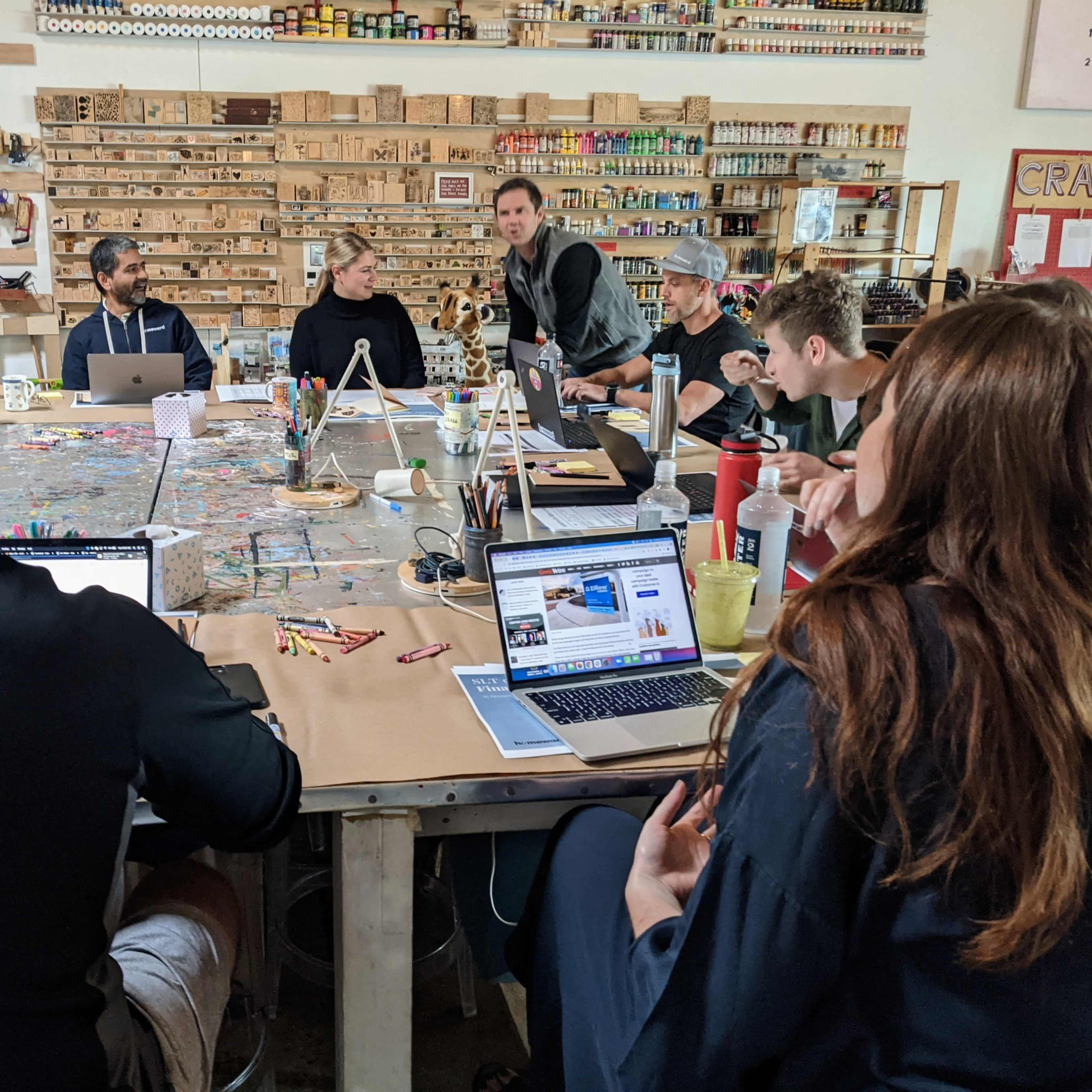 A group of people sits around a long table in a workspace, engaged in discussion while using laptops and other materials.