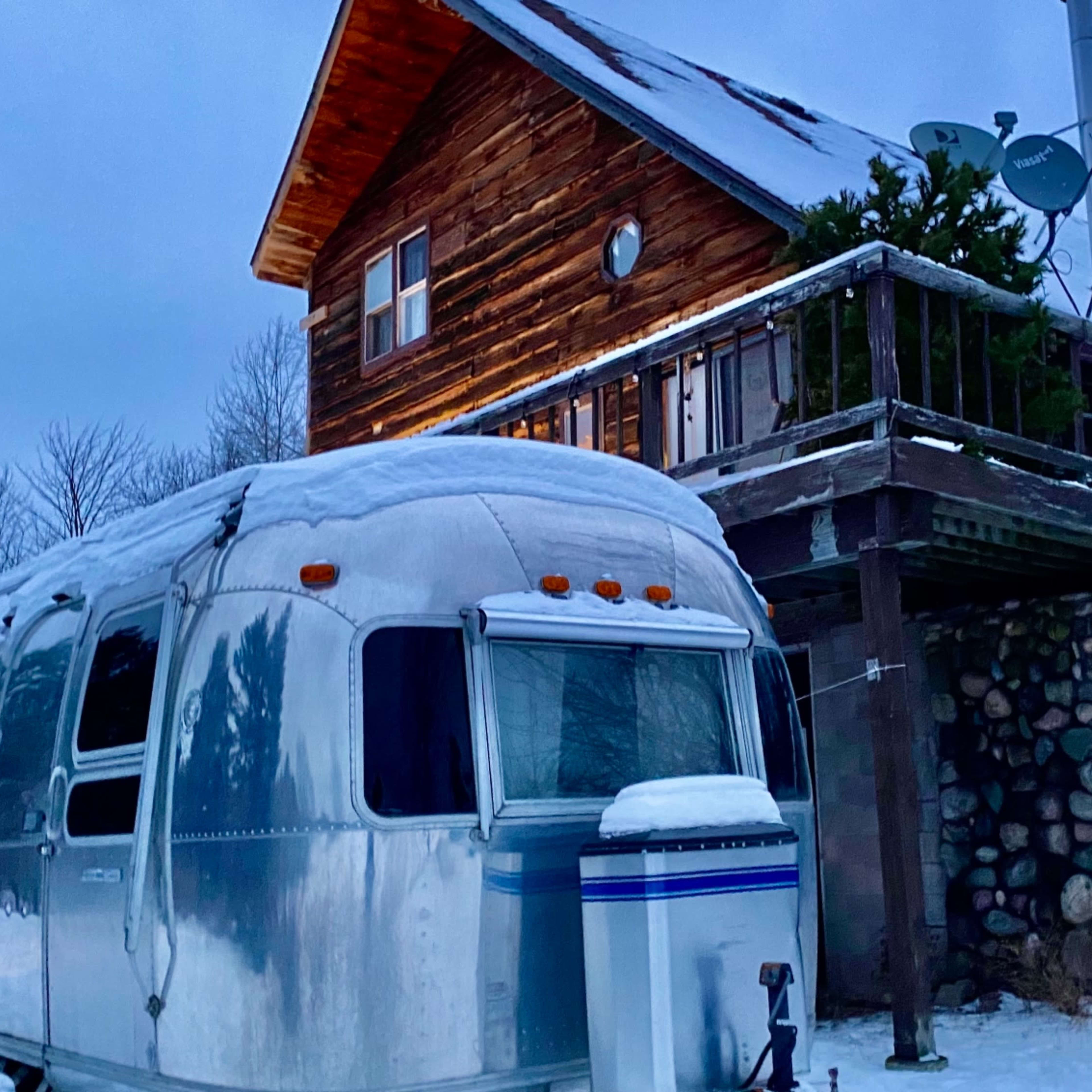 An Airstream trailer parked in a snow-covered landscape next to a wooden house with a balcony and satellite dishes.