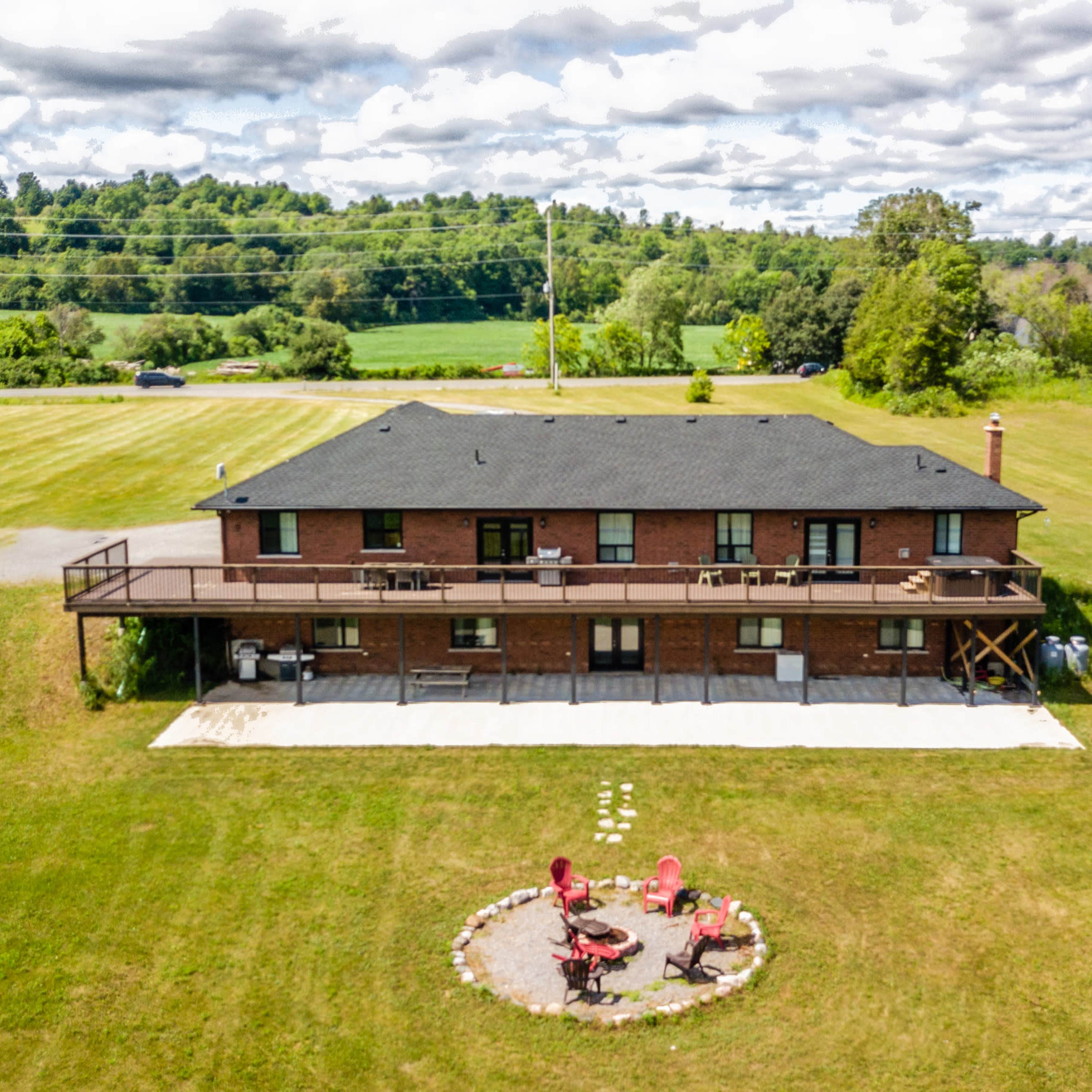 A large brick house with a wooden deck overlooks a green field, featuring a circular seating area with red chairs on the lawn.