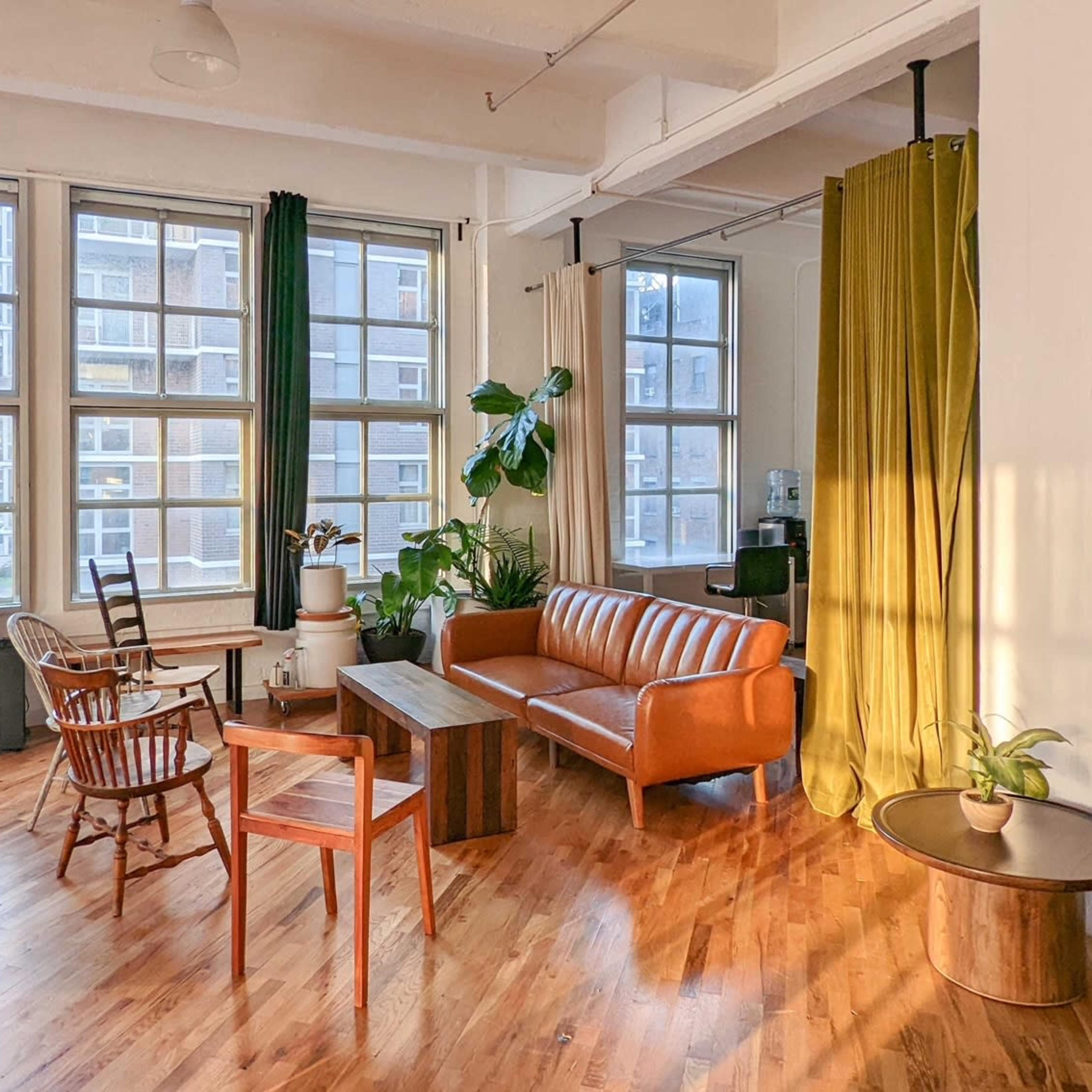 A bright, spacious room with large windows, featuring a brown leather sofa, wooden coffee table, and plants, along with a mix of green and gold curtains.