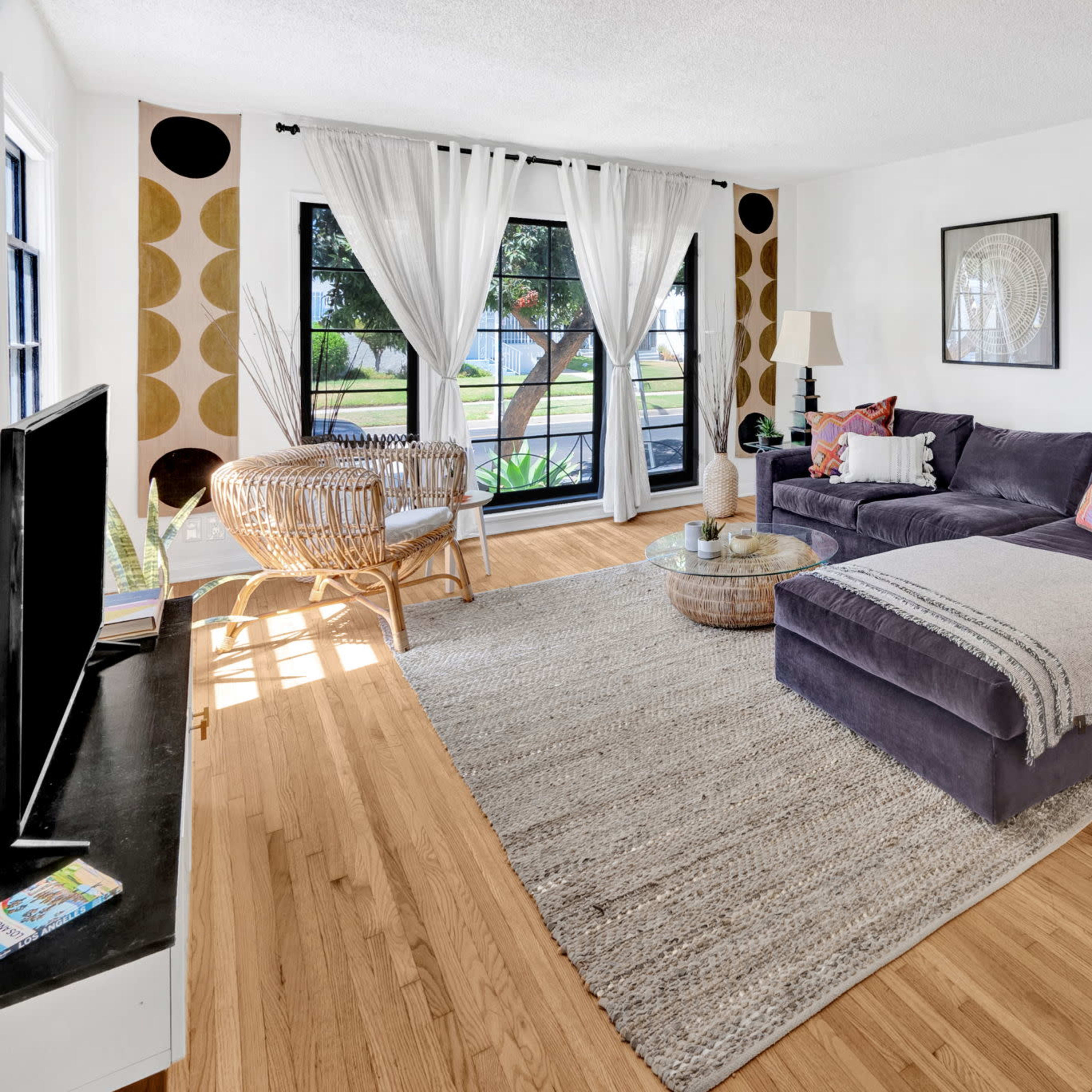 The living room features a grey sectional sofa, light hardwood flooring, large windows with sheer curtains, and a round coffee table in front of a television.
