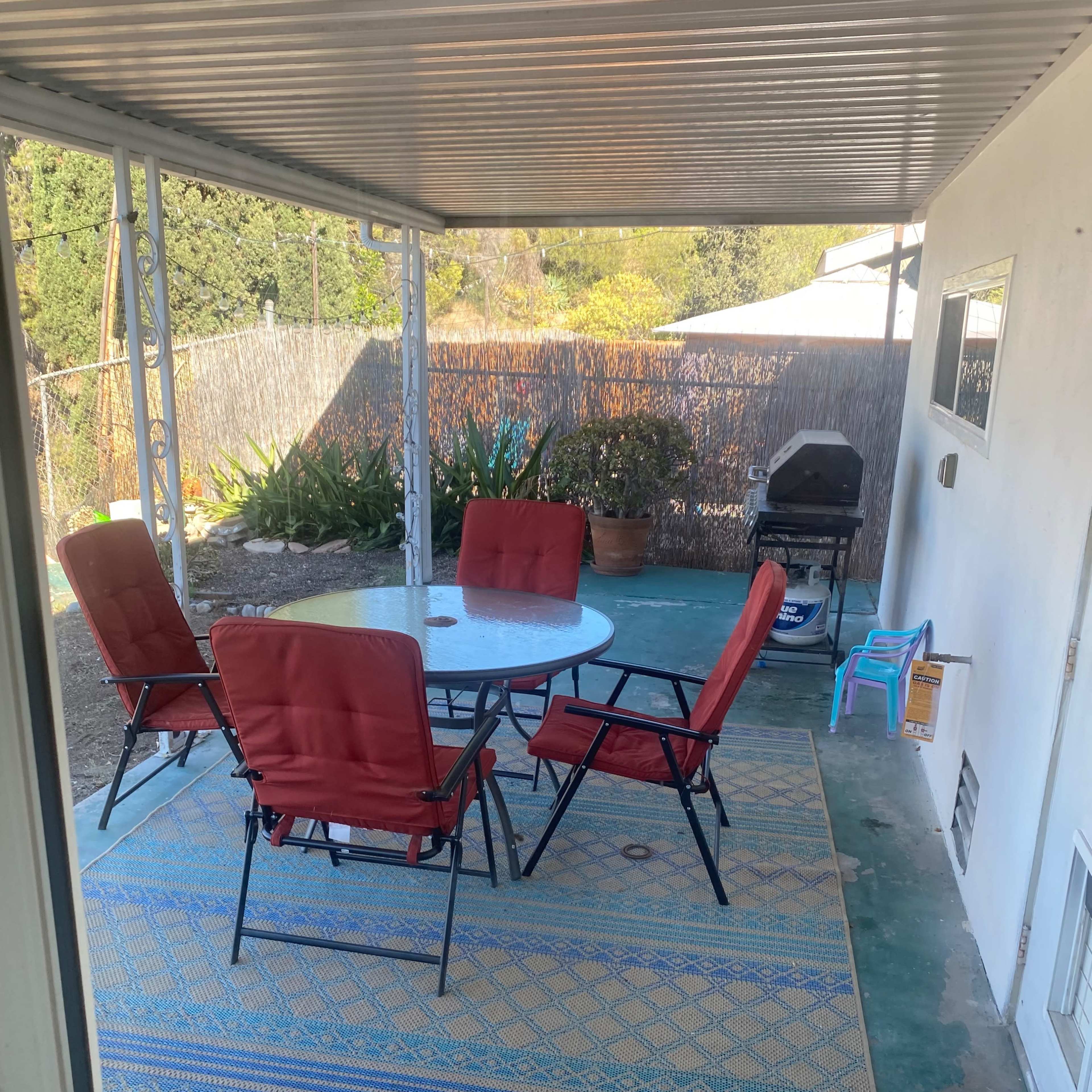 A covered patio features a round table with four red chairs, surrounded by potted plants and a barbecue grill in the background.