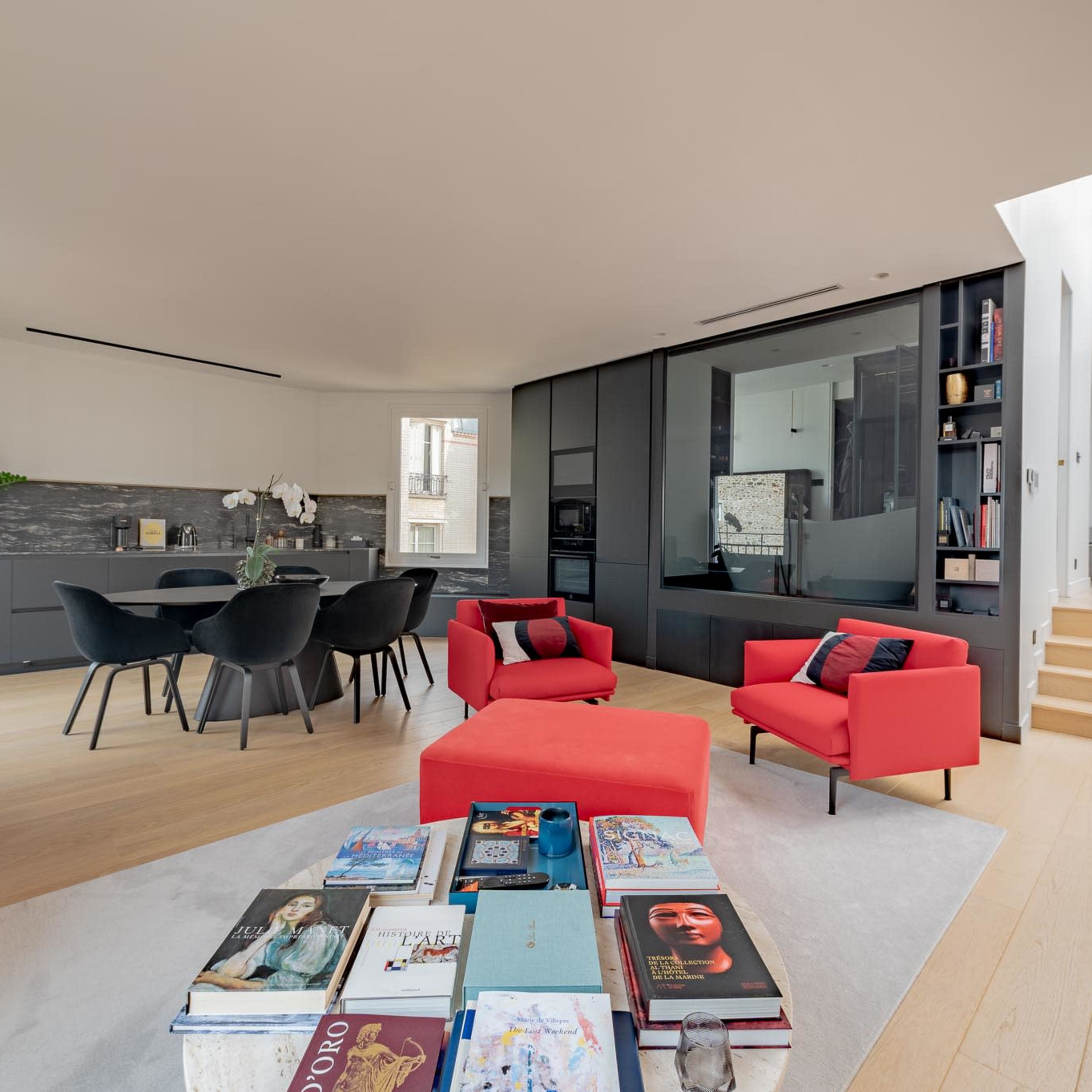 A modern living space features a dining area with a black kitchen, a seating arrangement of red chairs, and a coffee table stacked with books.