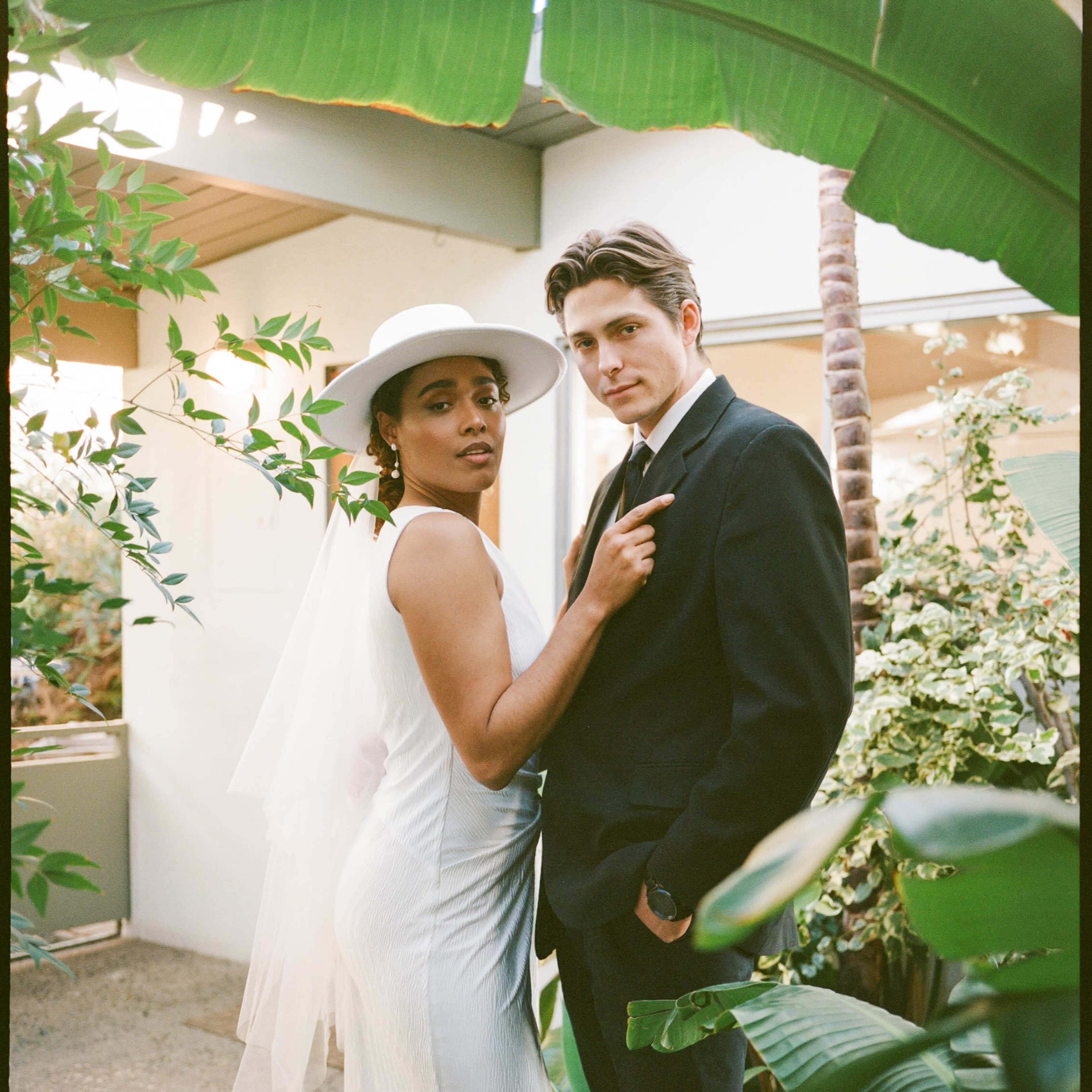 A bride in a white dress and a groom in a black suit pose together in a lush indoor garden setting.