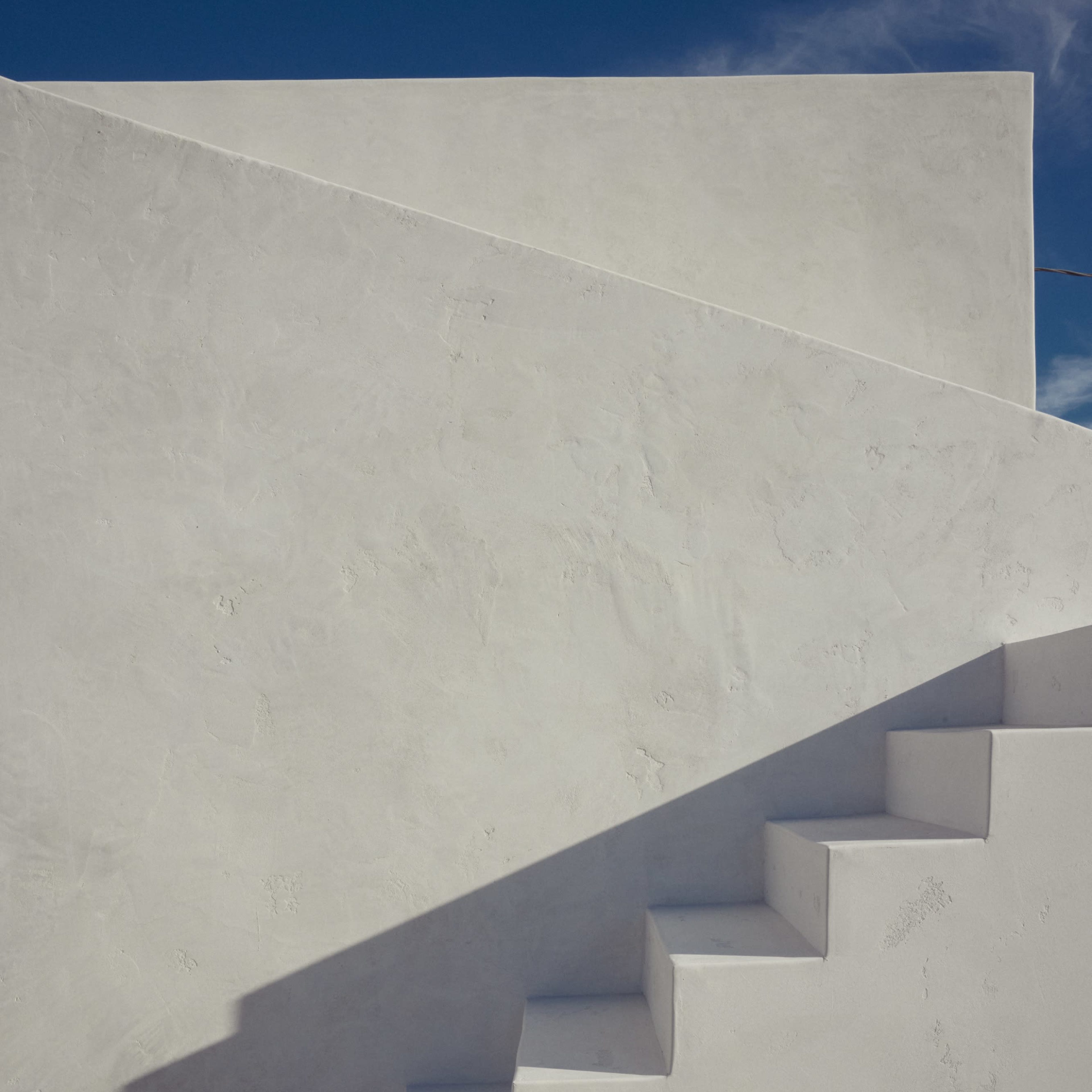 A series of white stairs ascend against a plain, textured wall under a clear blue sky.