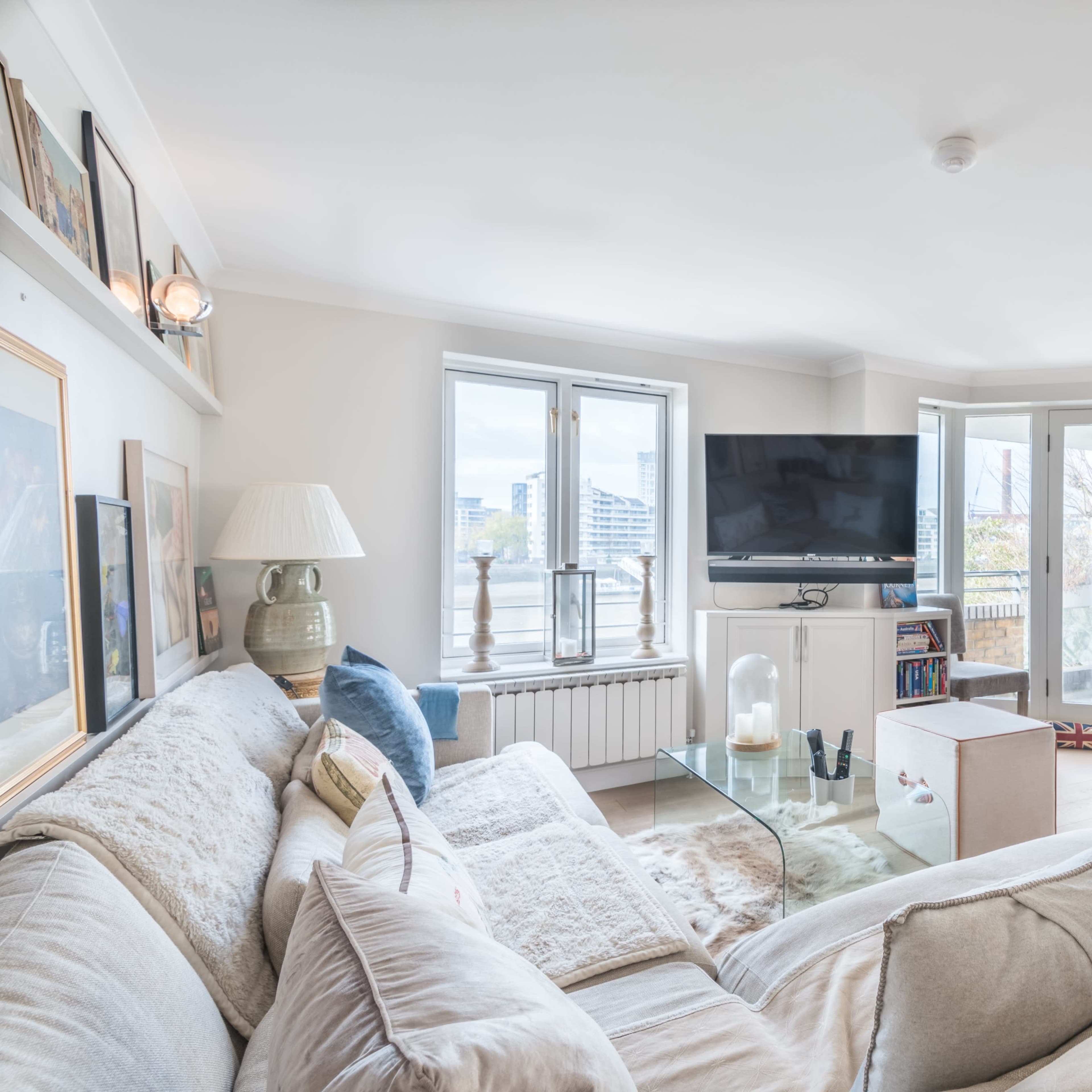 A modern living room features a large beige sectional sofa, a glass coffee table, and a flat-screen TV, with large windows that offer views of the outdoors.