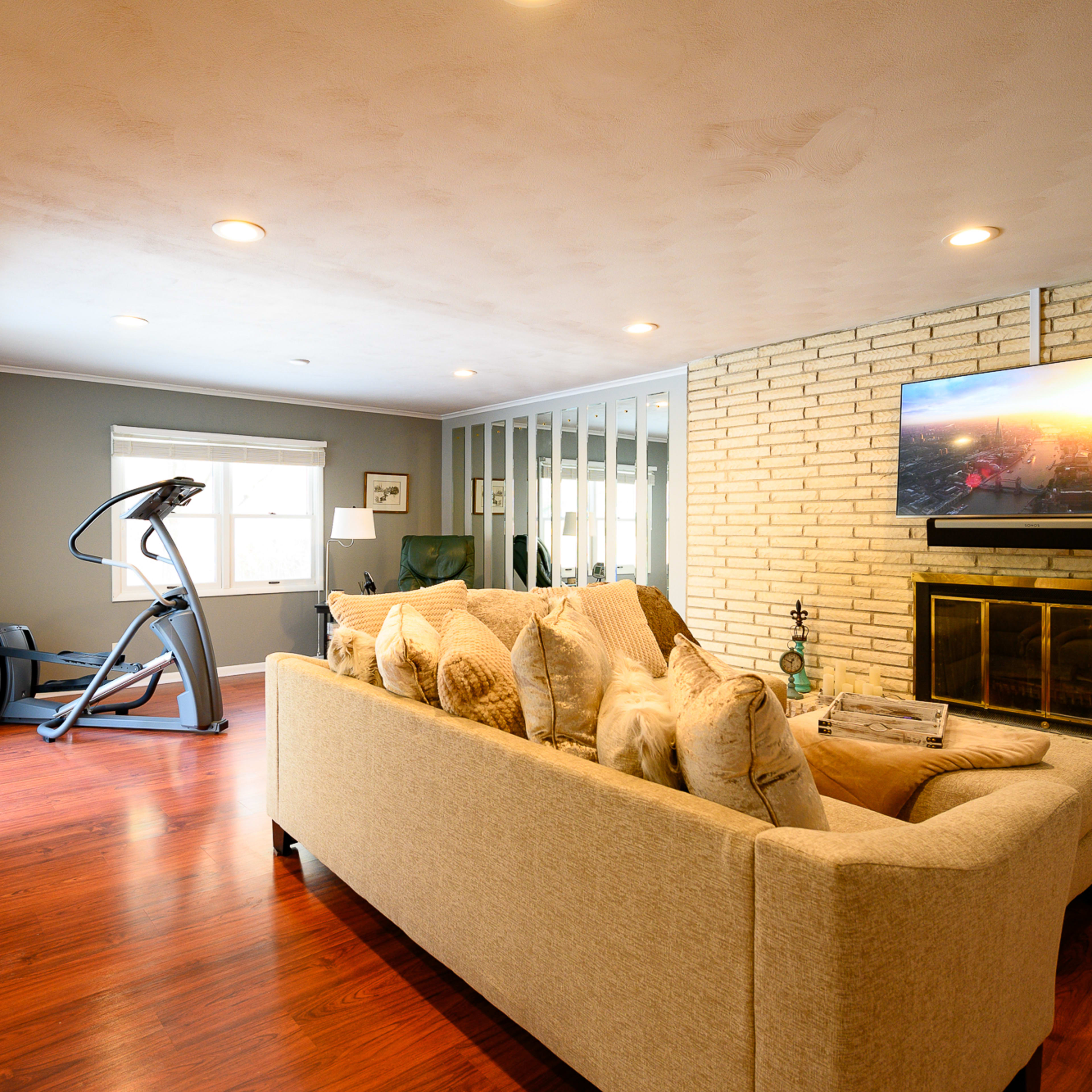 A living room with a beige couch, a television mounted above a brick fireplace, and an exercise machine near a window.