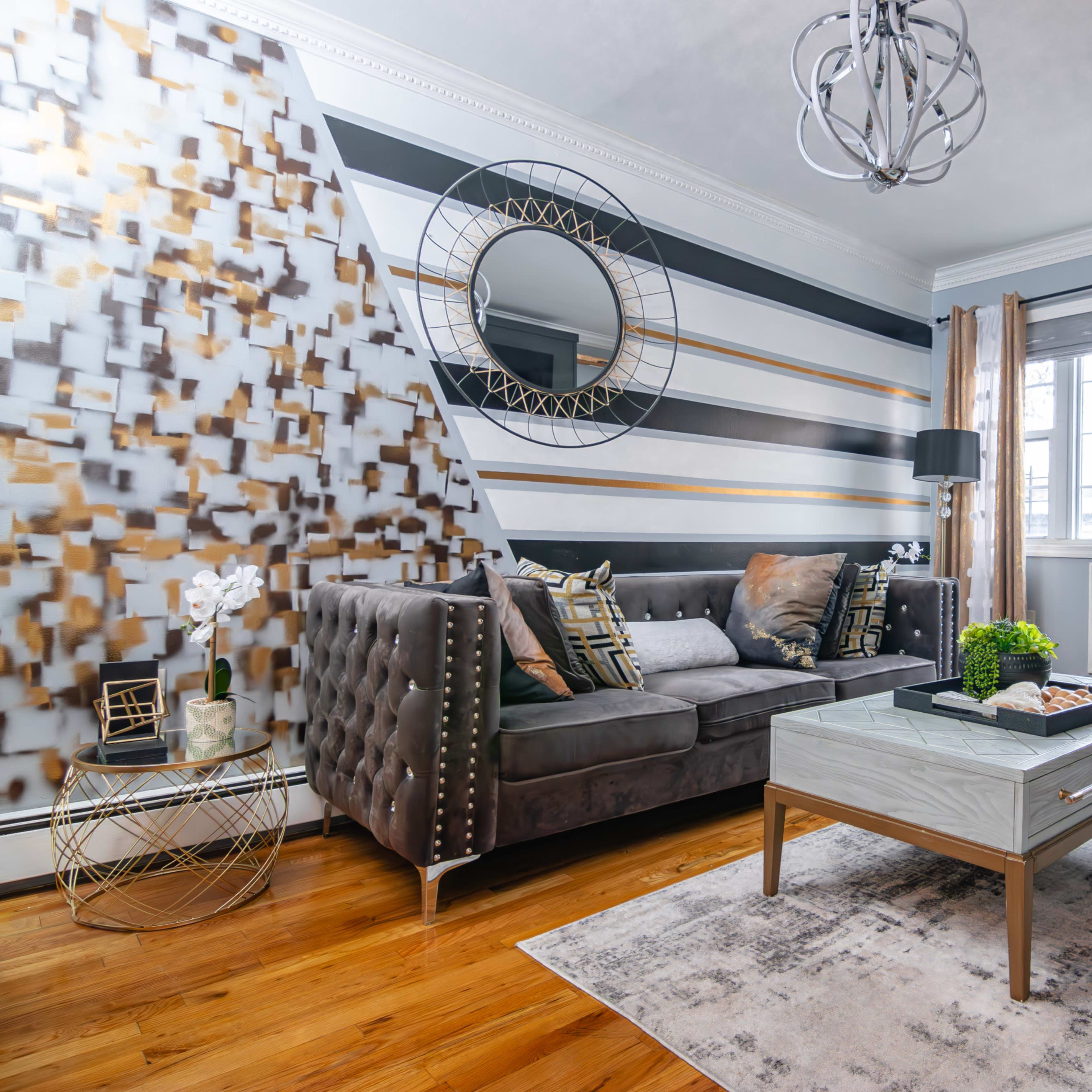 A modern living room with a tufted gray sofa, a mirrored coffee table, and a decorative wall combining geometric patterns in black, white, and gold.