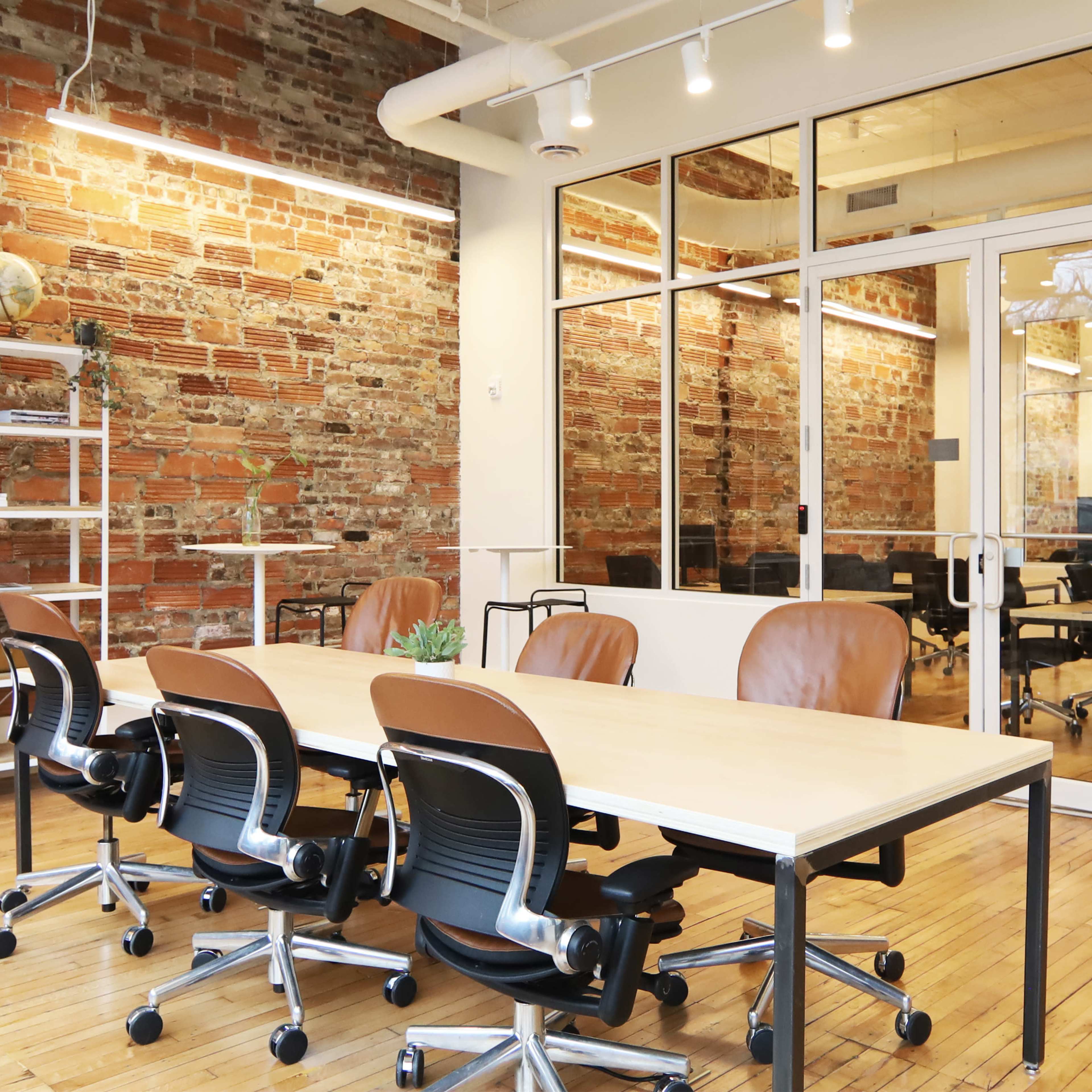 A modern office space with a long table and several ergonomic chairs, featuring exposed brick walls and large windows.
