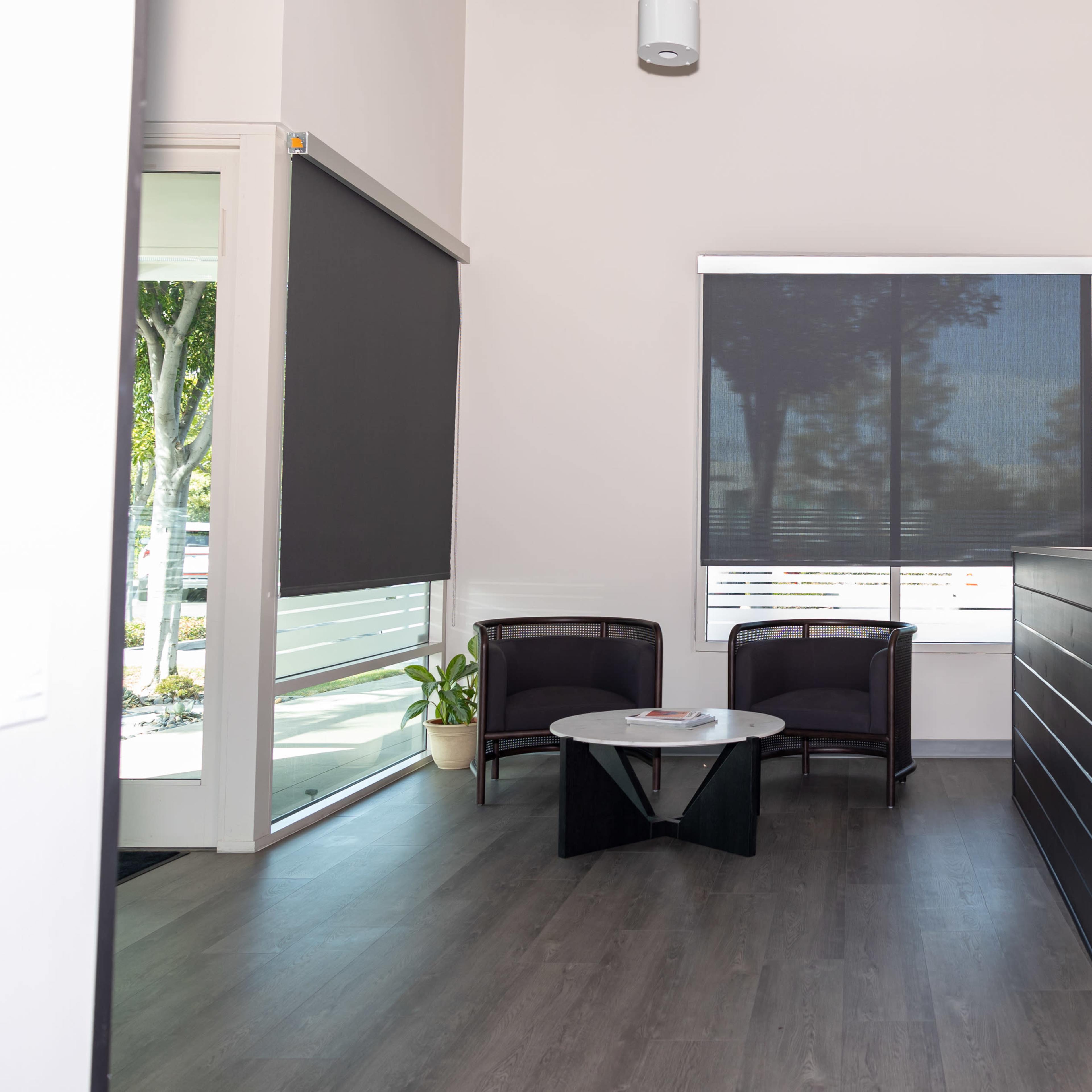 A modern reception area with two chairs, a small table, and a white front desk next to large windows covered by dark blinds.