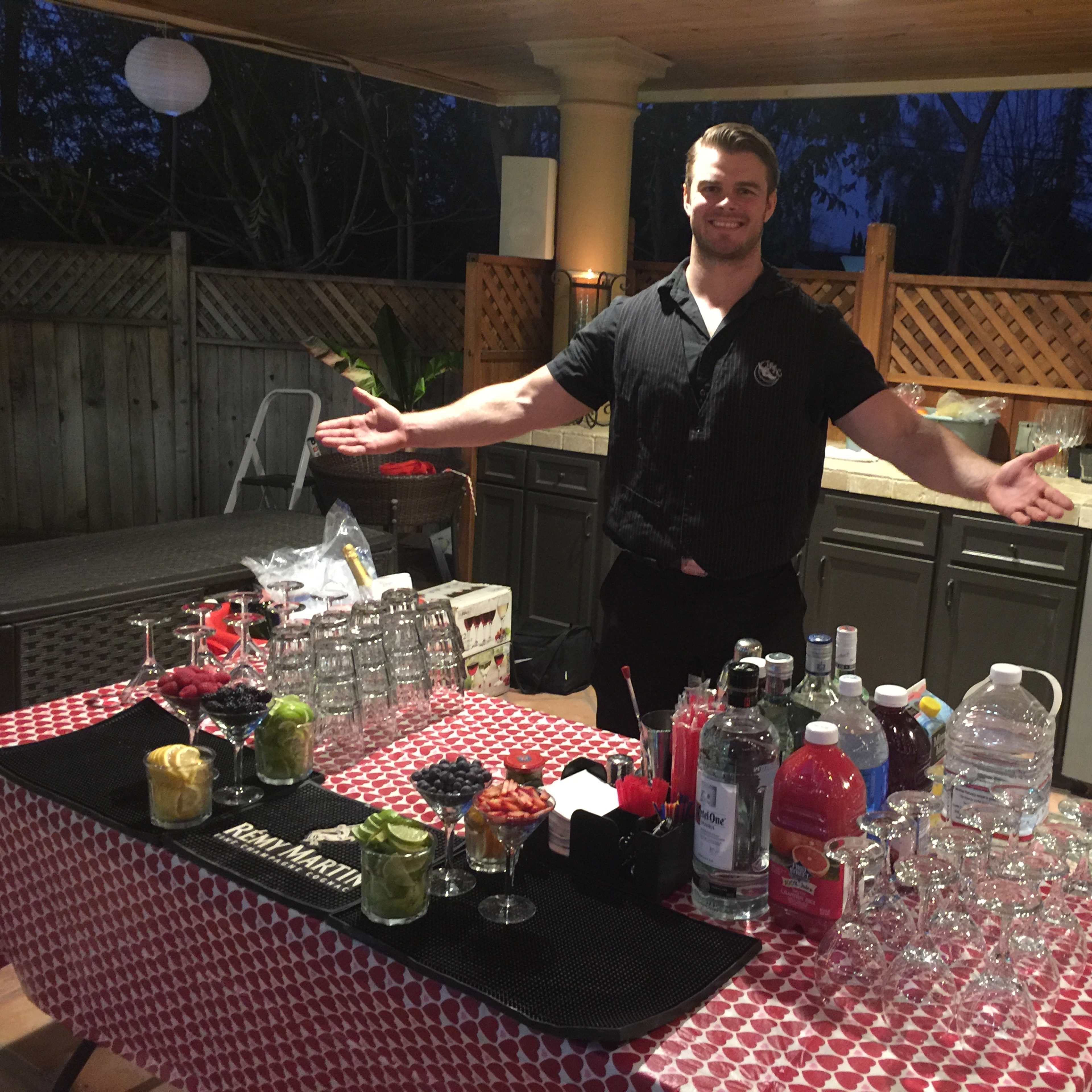 A bartender stands behind a table filled with various bottles and colorful ingredients for making cocktails in a backyard setting.