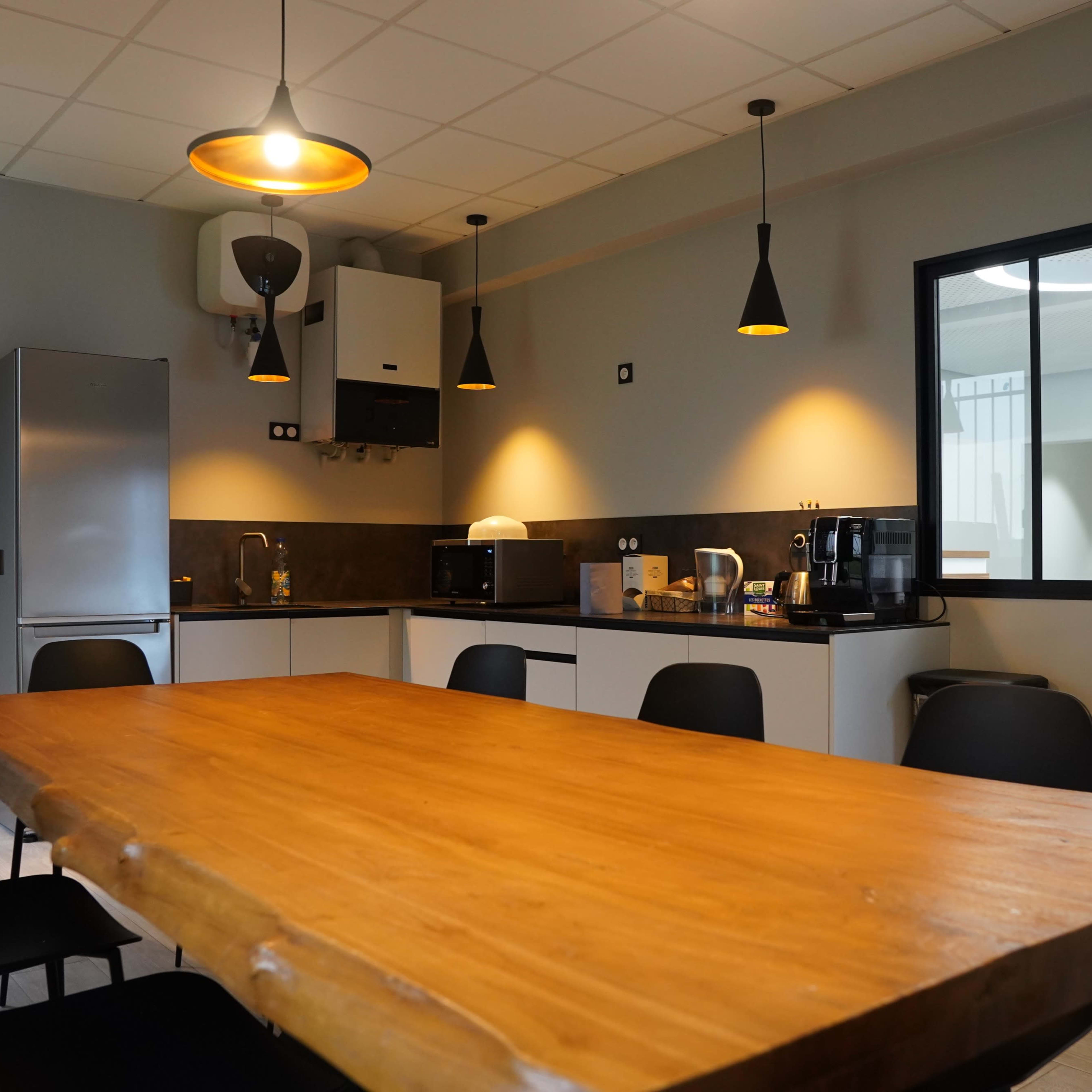 A modern kitchen with a large wooden table, black chairs, and various appliances including a fridge and coffee maker.