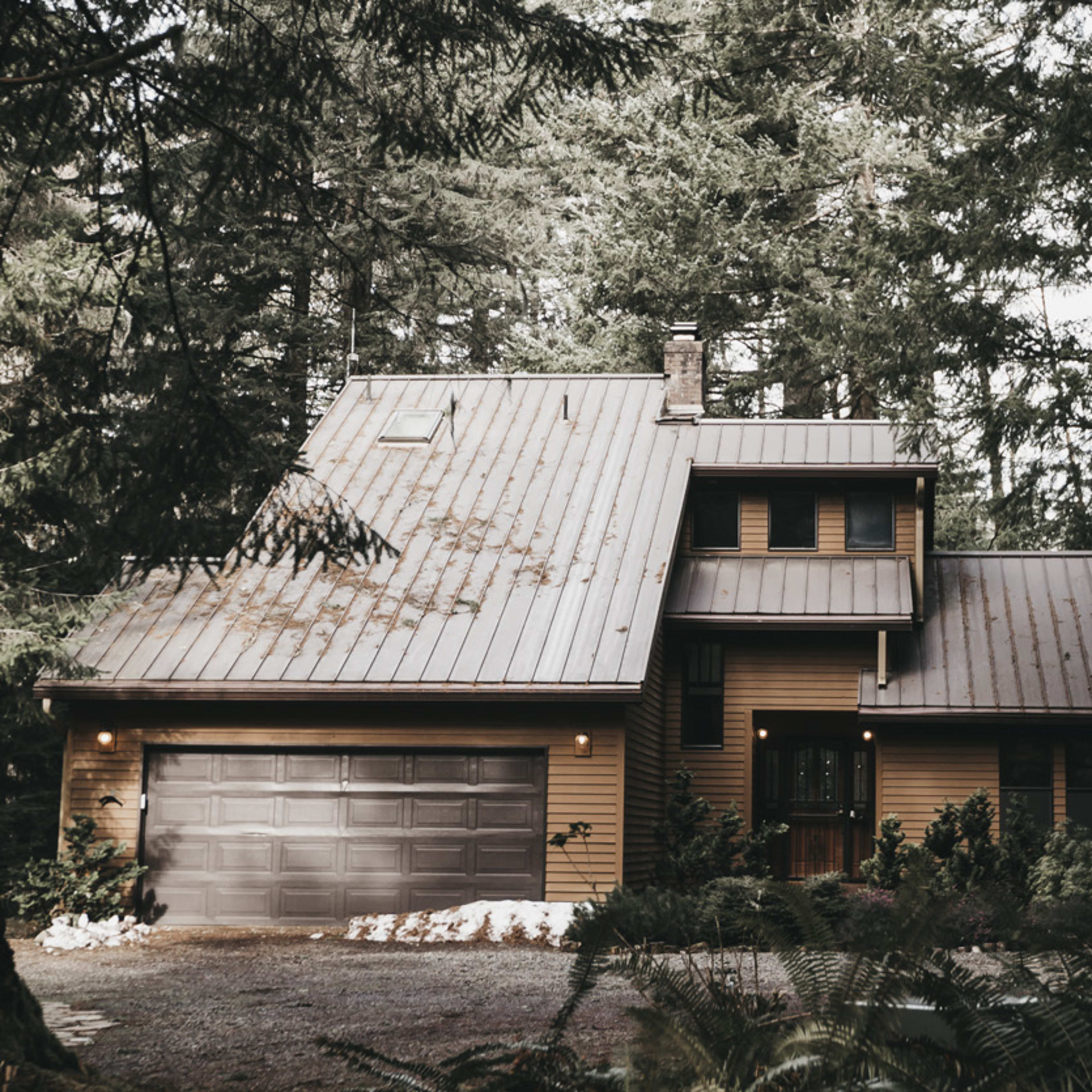 A two-story wooden house with a metal roof is situated among tall trees in a forested area.
