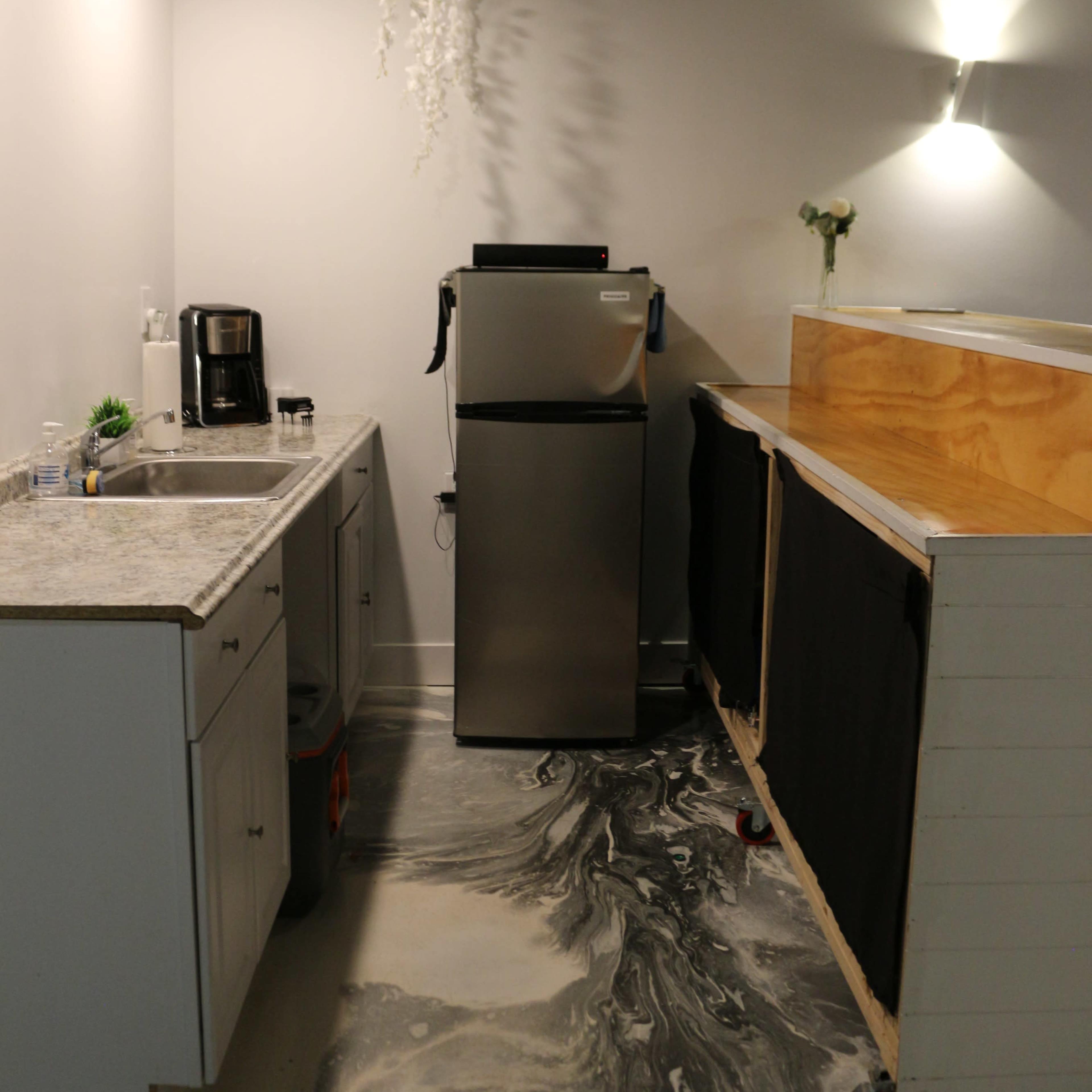 A kitchen area features a countertop with a coffee maker, a silver refrigerator, and a wooden bar with black fabric curtains, all set on a marbled floor.