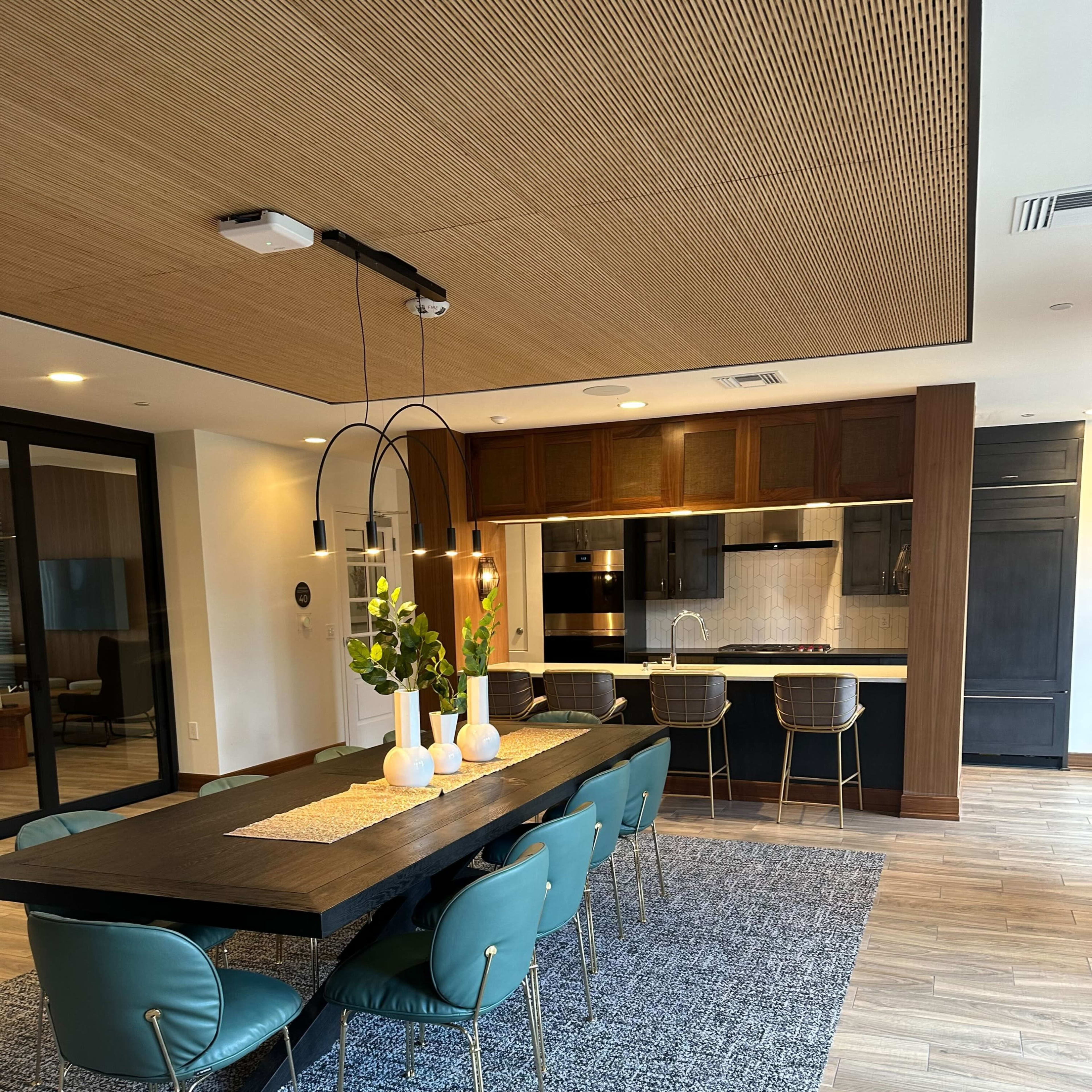 A modern dining area with a large rectangular table, teal chairs, and a view of a kitchen with dark cabinetry and stylish appliances.