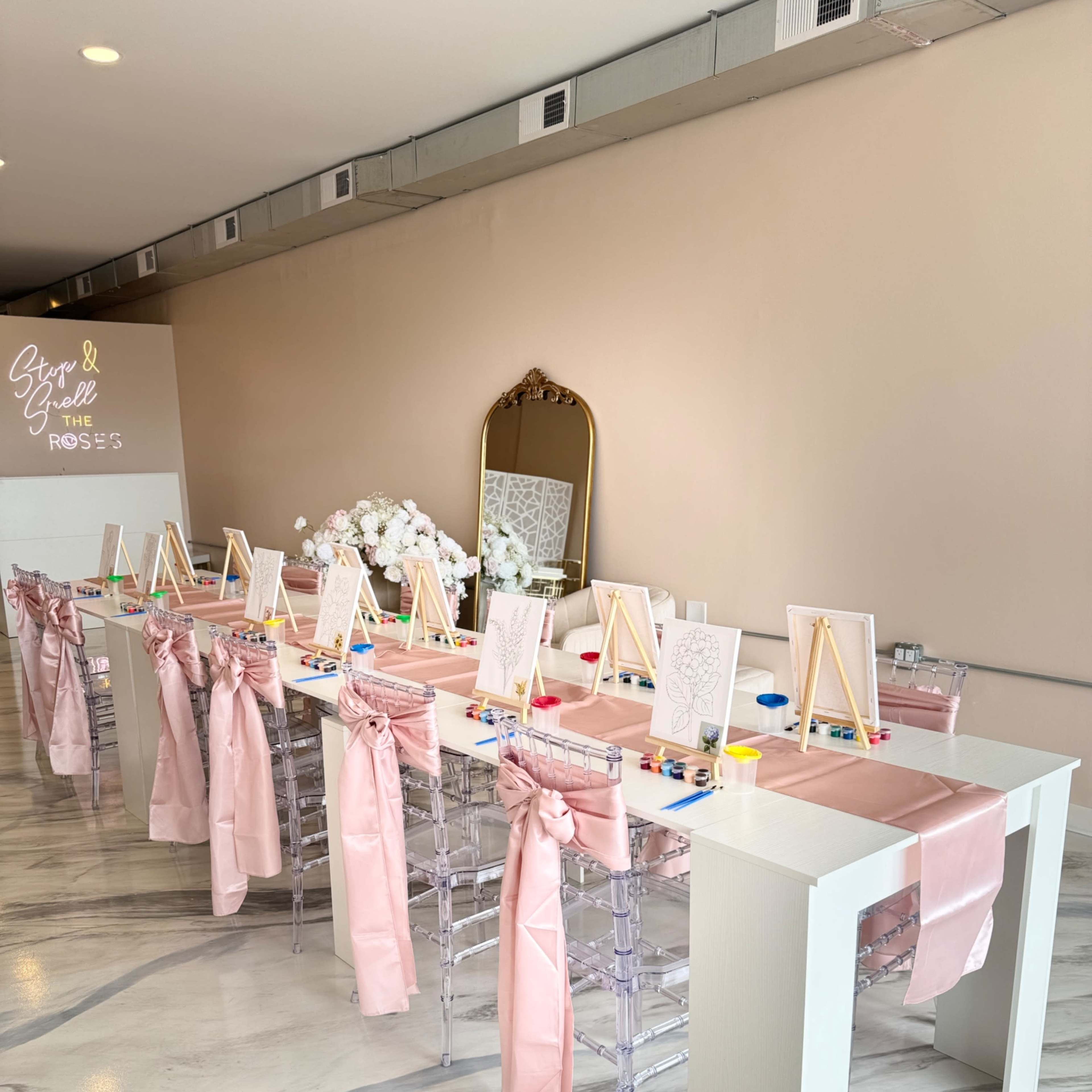A long table is set for an art activity, featuring pink sashes on clear chairs, small easels, and art supplies arranged along the surface.