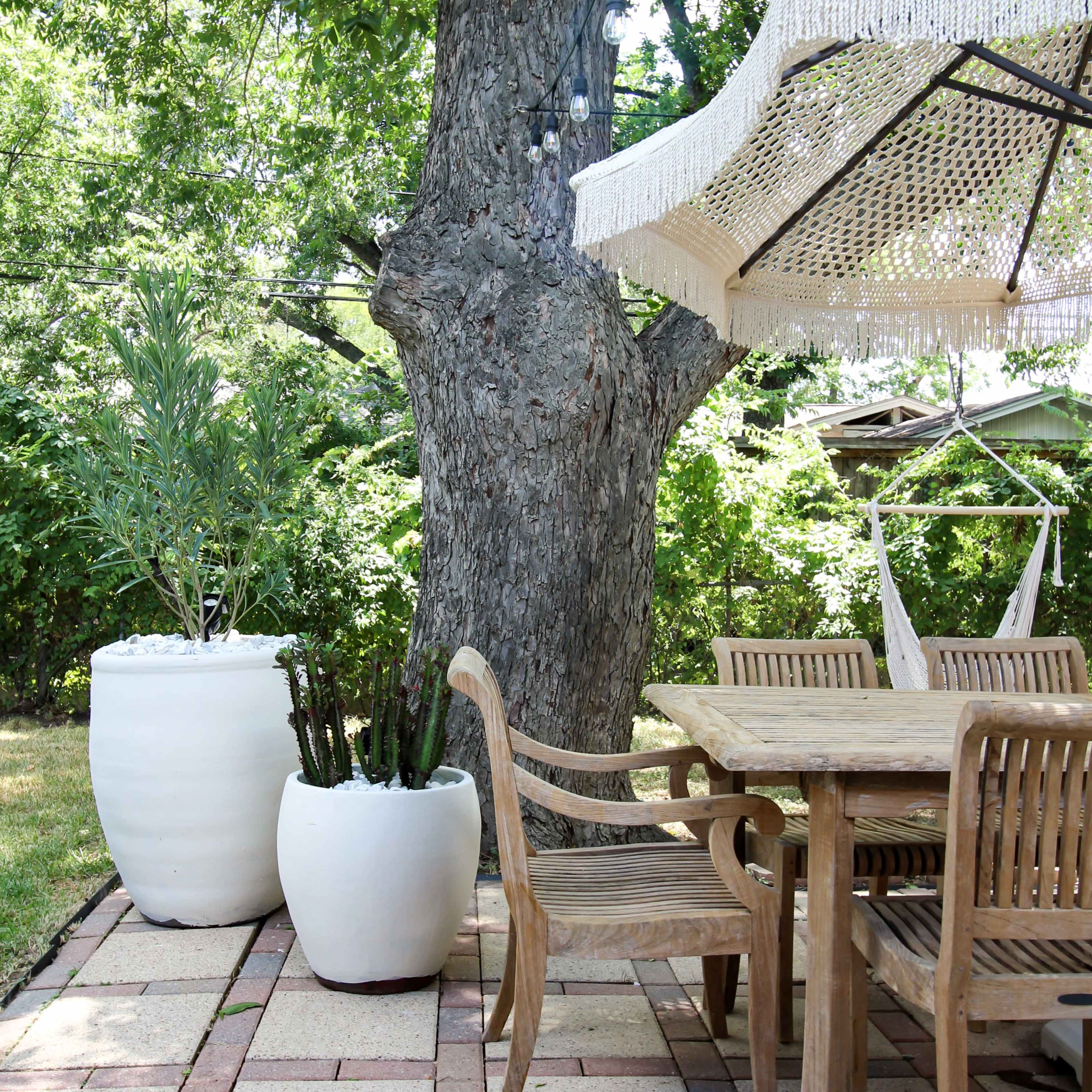 A wooden dining table and chairs under a patio umbrella beside large white planters and a tree in a garden setting.