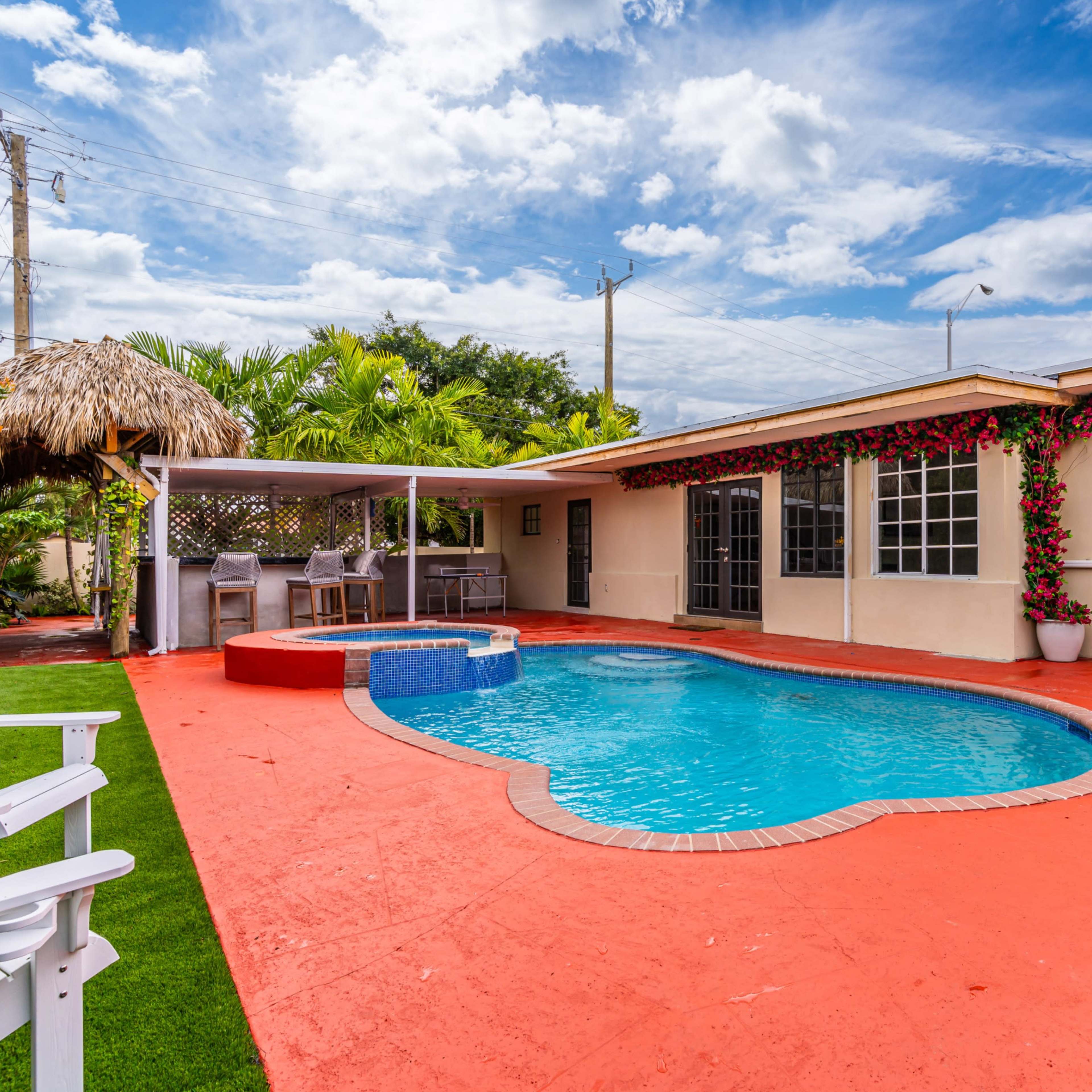 A backyard with a swimming pool surrounded by red-painted concrete, a thatched gazebo, and lush greenery.