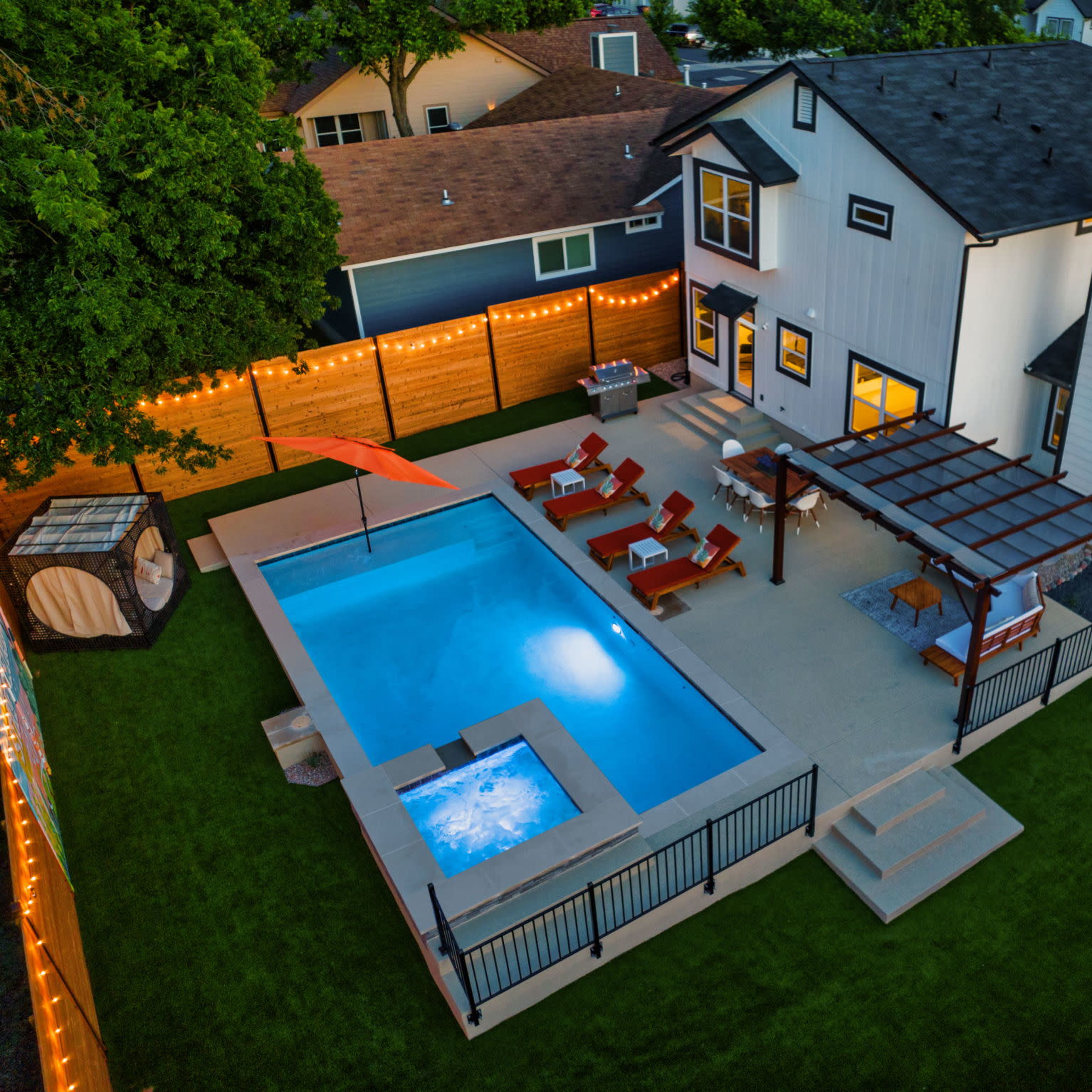A backyard featuring a swimming pool, a hot tub, lounge chairs, and a pergola, all surrounded by wooden fencing and string lights.