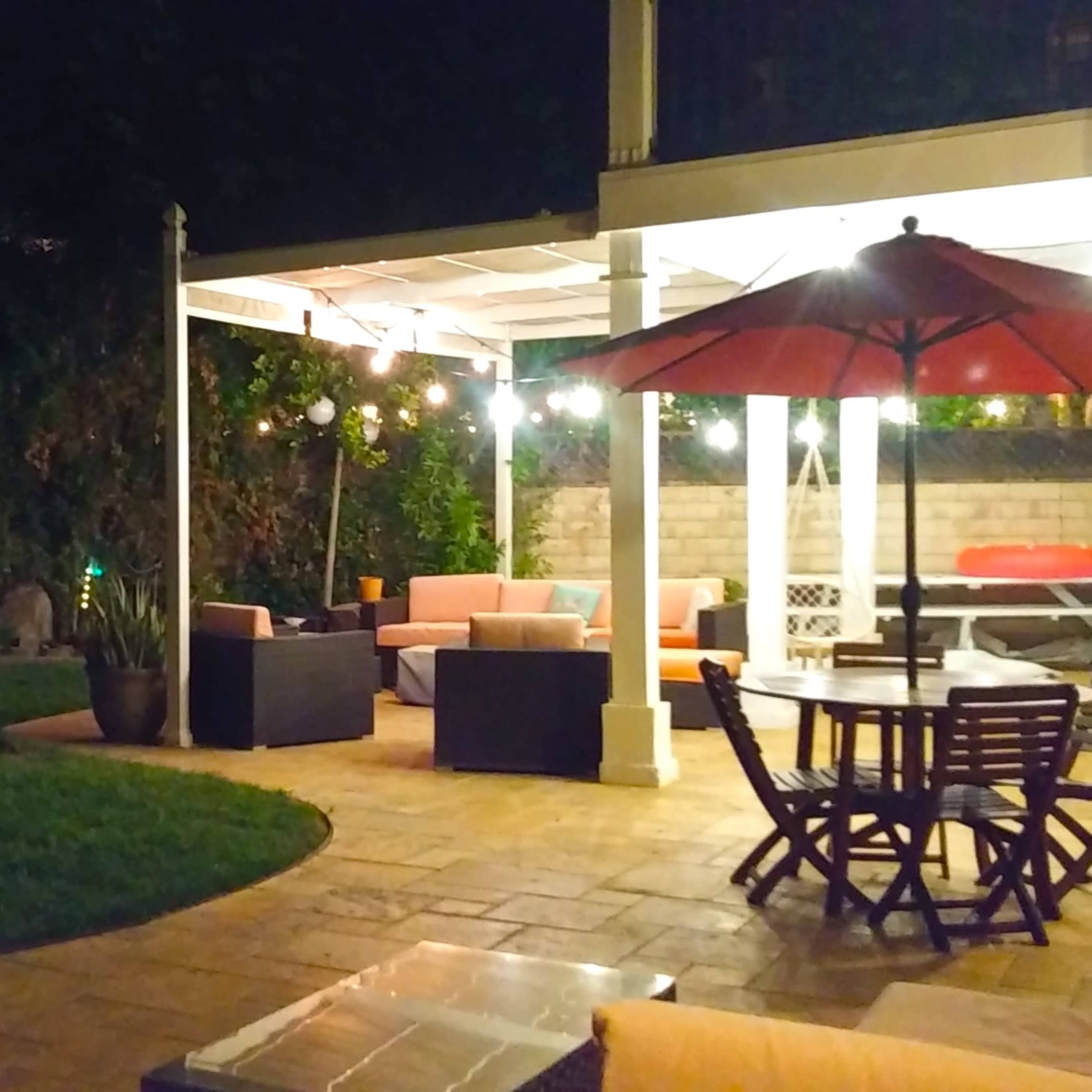A well-lit backyard features a seating area with couches, a dining table, and decorative string lights under a covered patio.