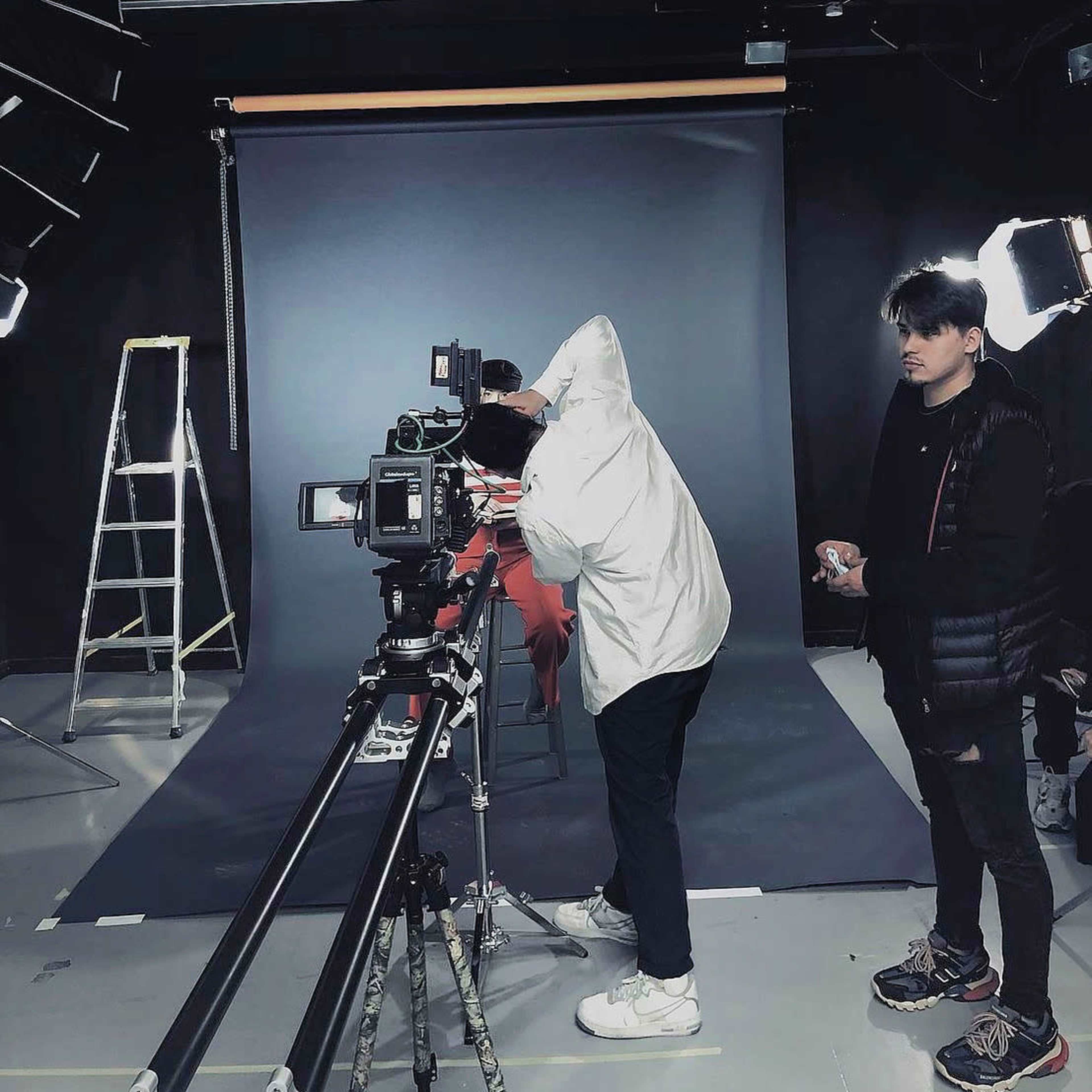 A person in a white jacket operates a camera on a tracking dolly while another individual stands nearby in a dimly lit studio with a backdrop and lighting equipment.