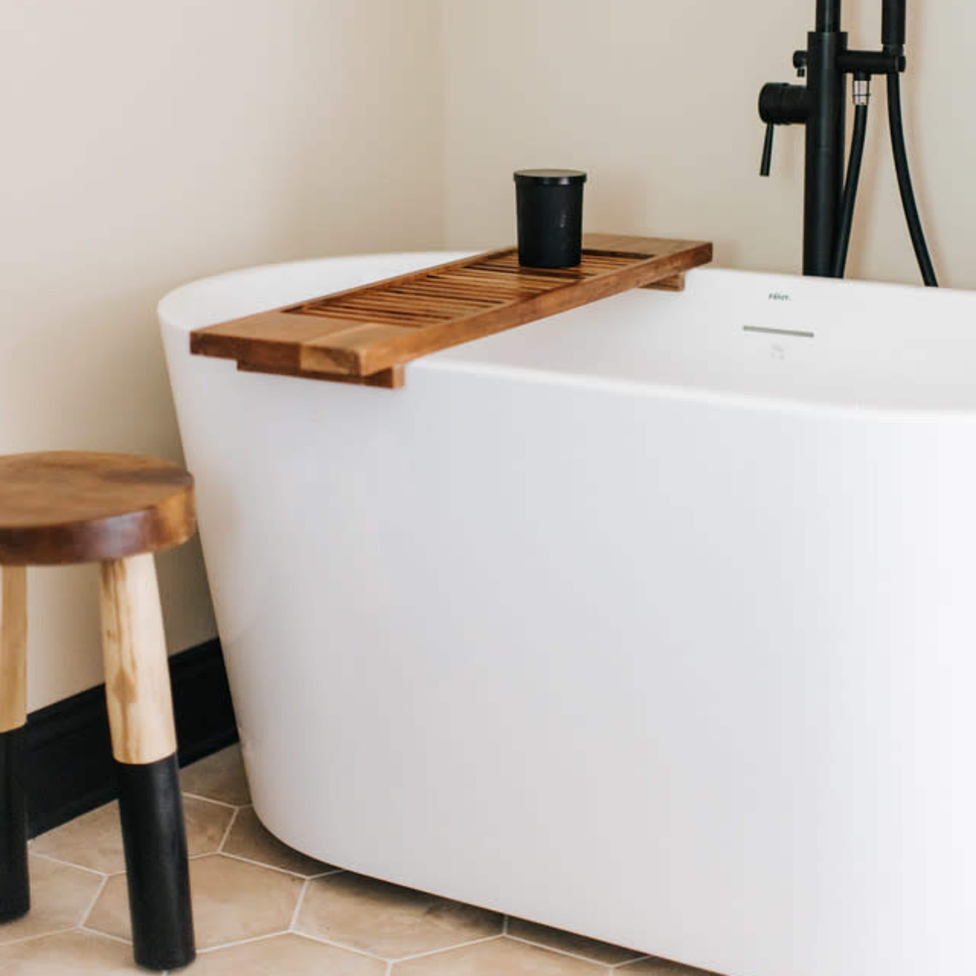 A freestanding white bathtub with a wooden bath caddy sits next to a small wooden stool against a light-colored wall.