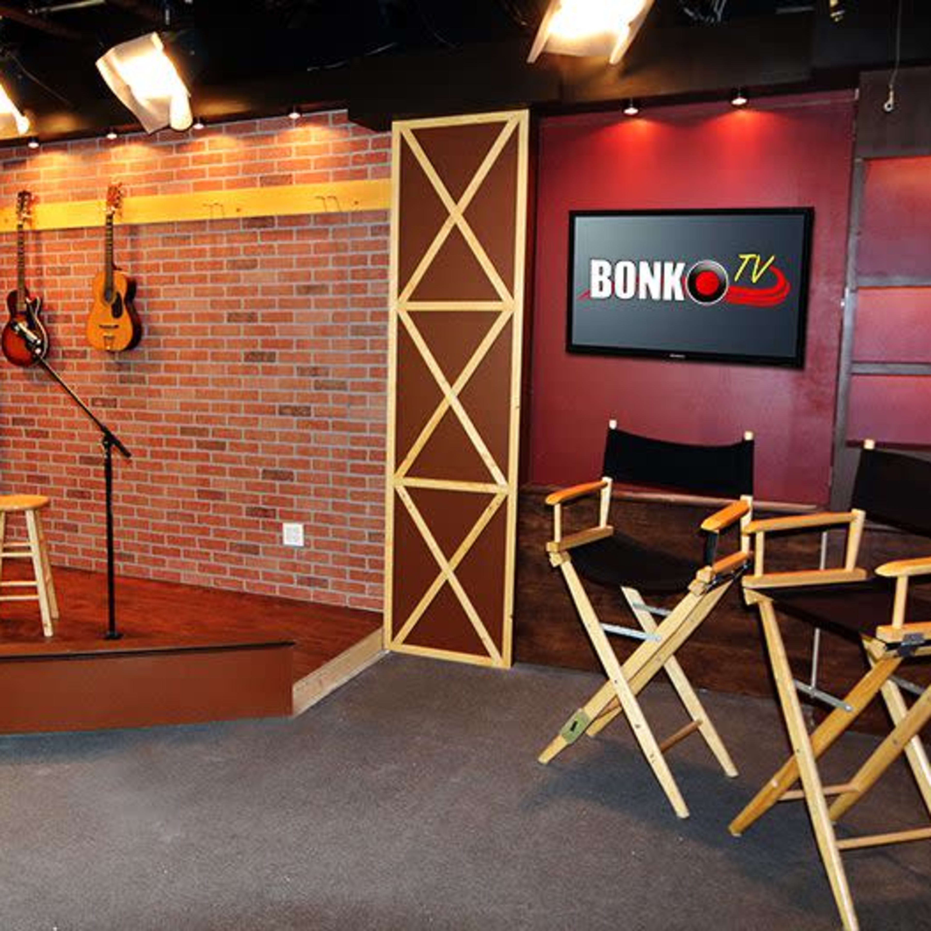 A television studio set featuring two microphone stands, two stools, and director's chairs against a brick backdrop with decorative lighting and guitars on display.