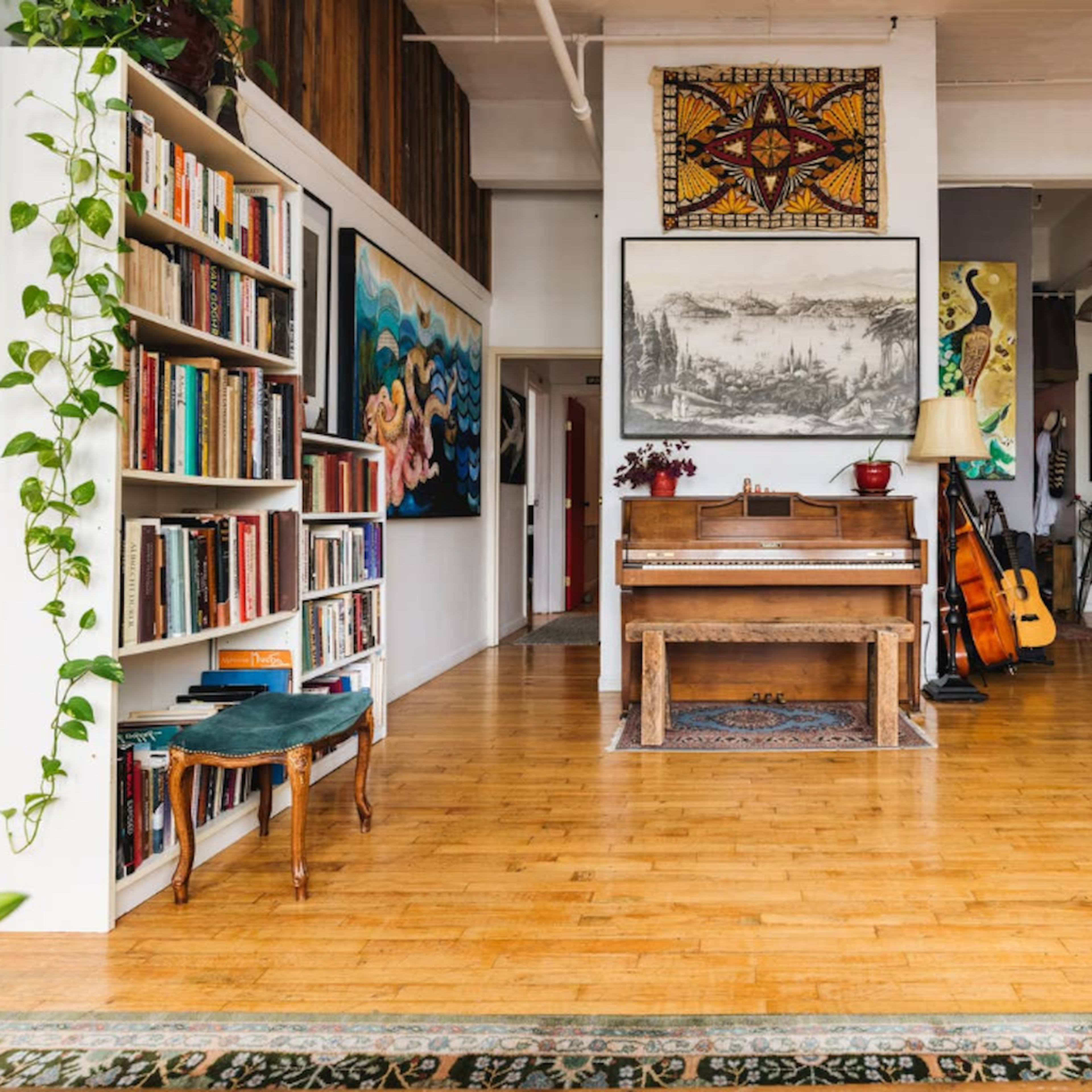 A spacious room with hardwood floors, featuring a piano, bookshelves filled with books, and various artistic decorations on the walls.