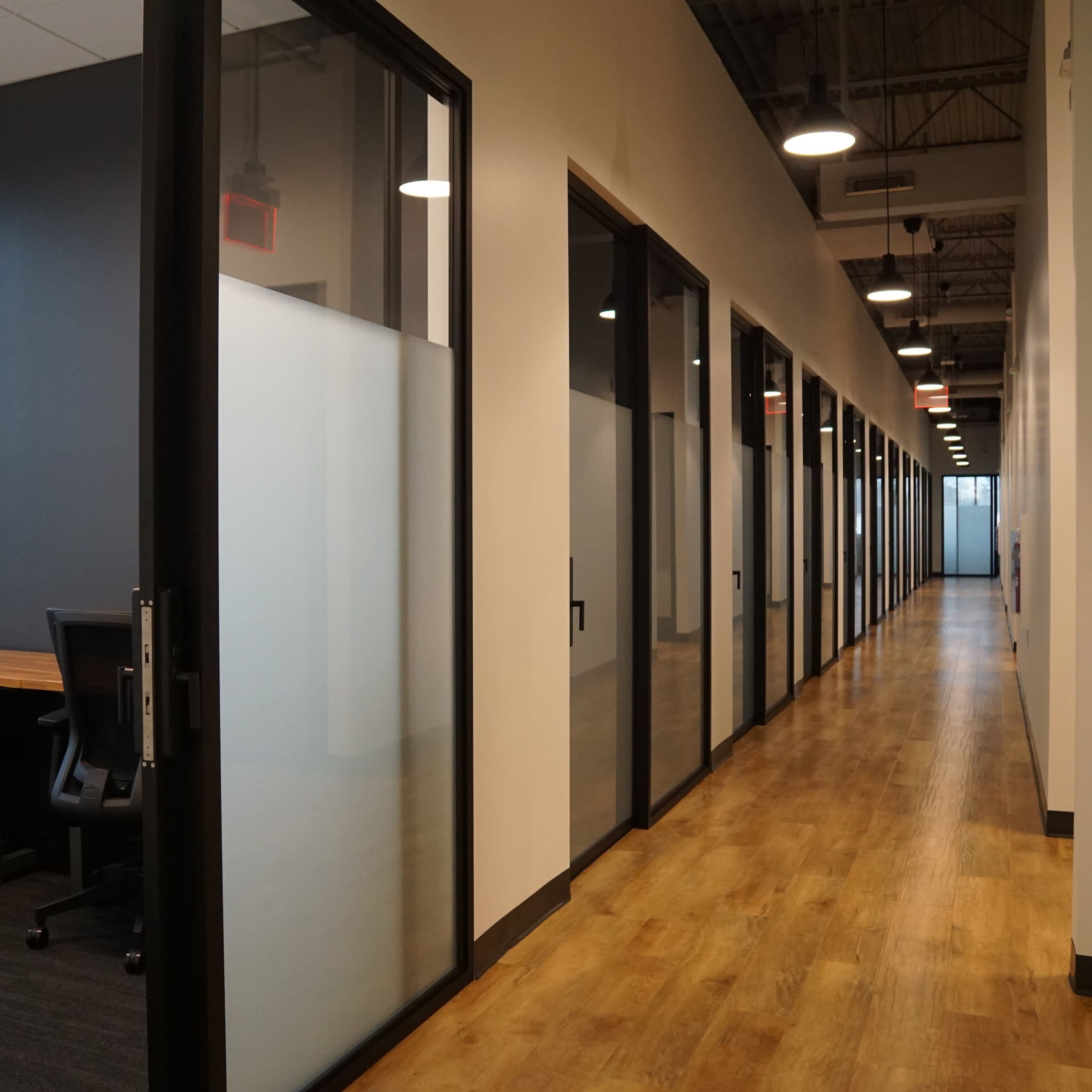 A modern office hallway features glass-walled meeting rooms on one side and a wooden floor leading to additional workspace.