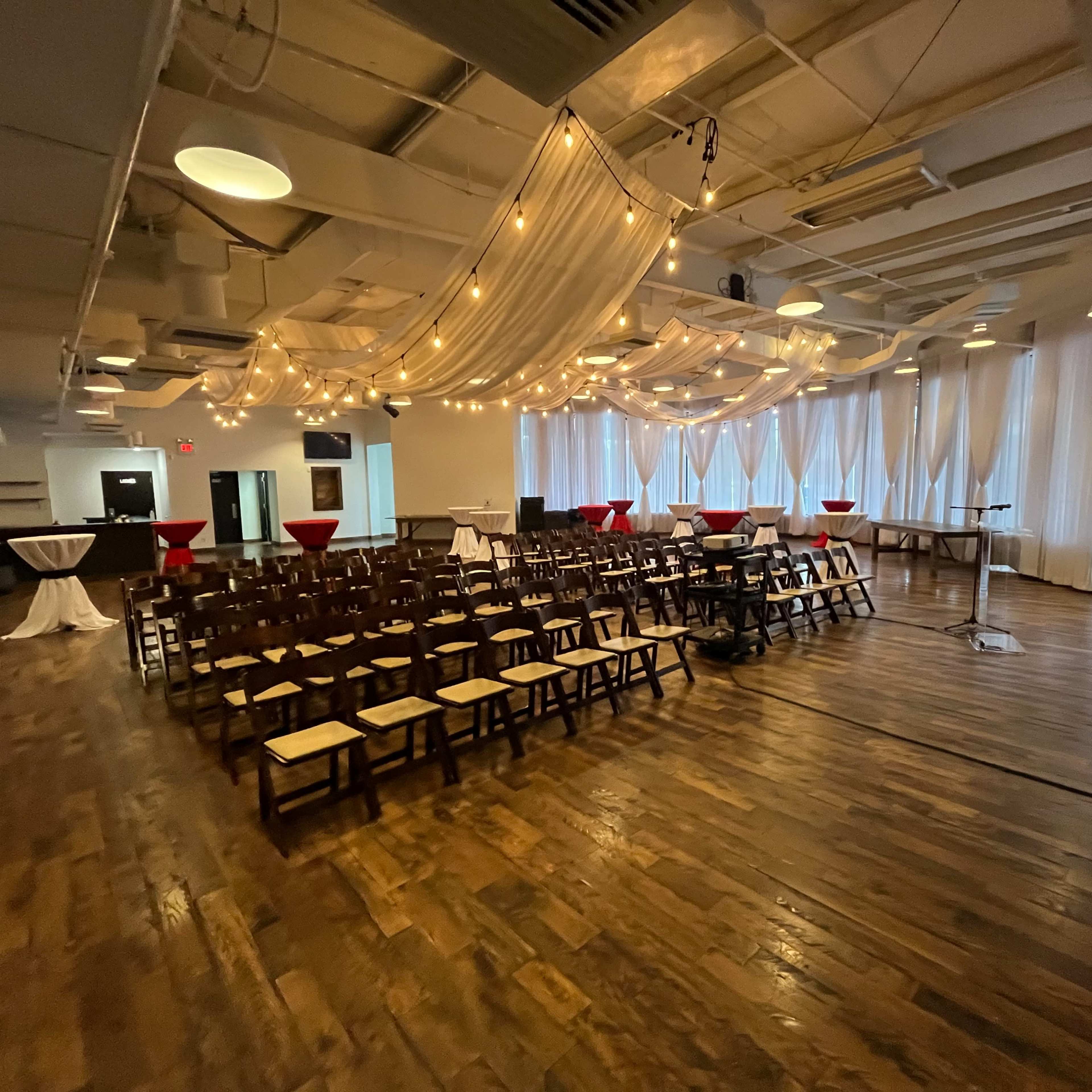 An event space set up for a gathering, featuring rows of wooden chairs, cocktail tables with red cloths, and decorative drapes overhead.