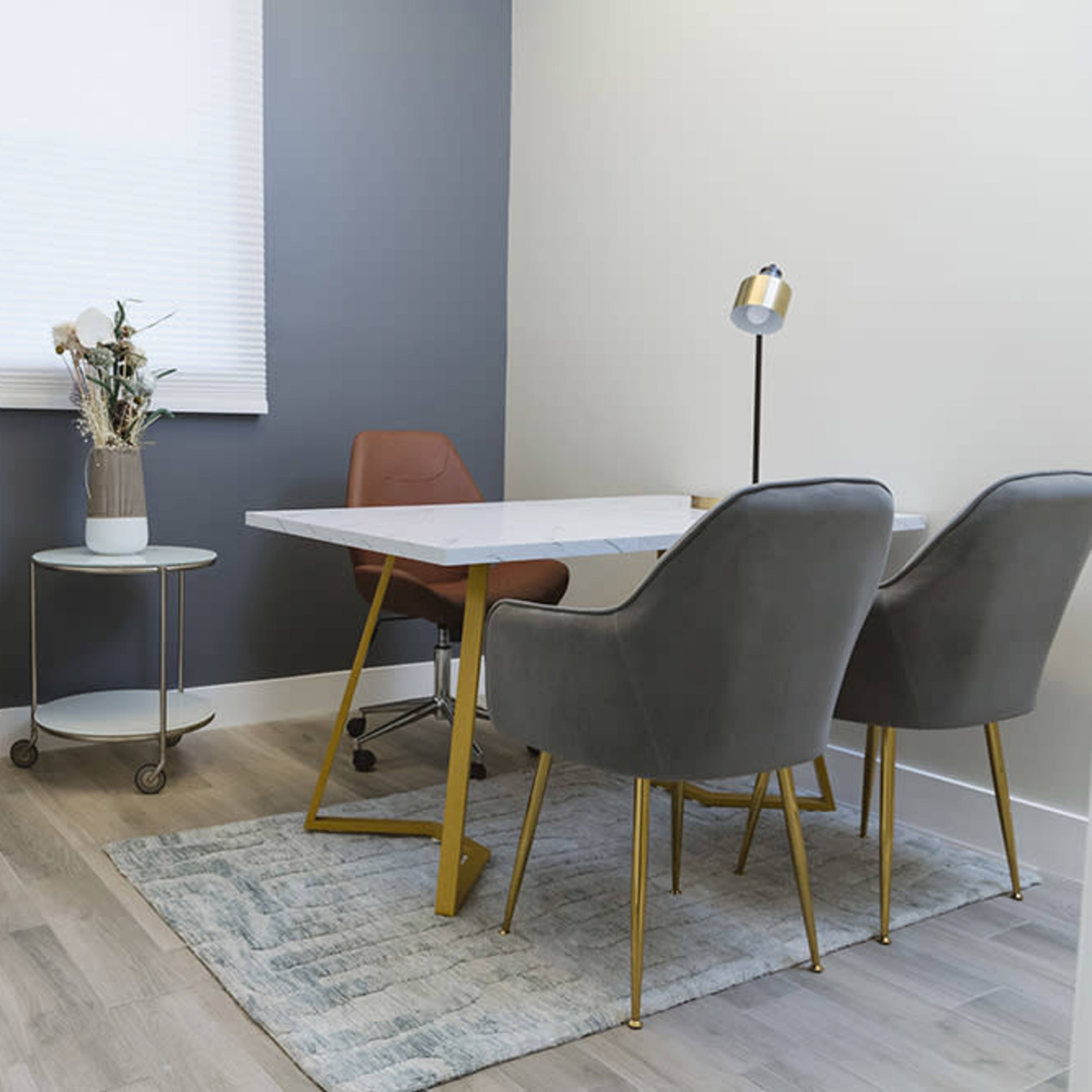 A small office with a round gray chair, a white table with gold legs, two gray chairs, a lamp, and a small side table featuring a vase with dried flowers.