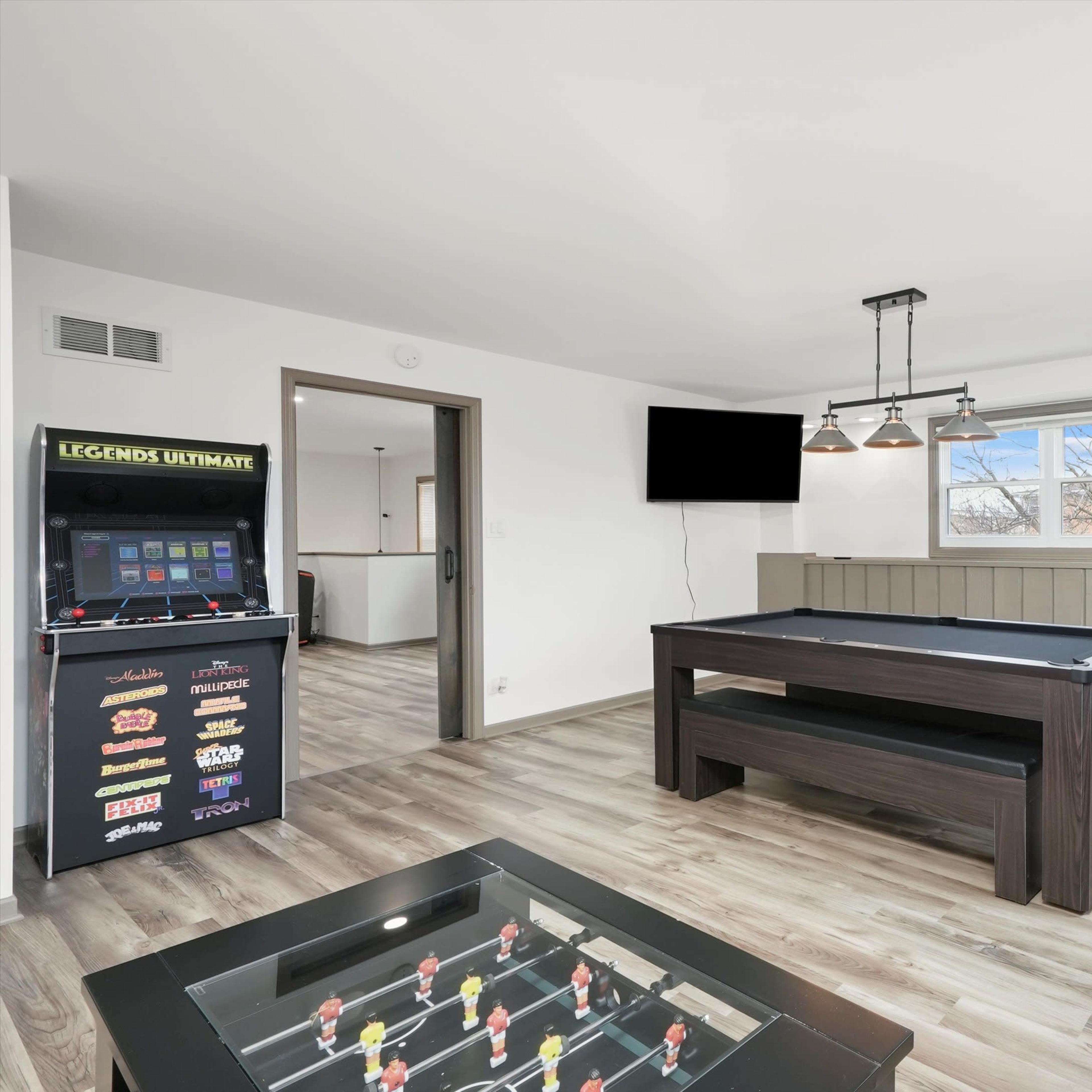 A modern game room featuring a pool table, arcade machine, and bar-height seating near a large window.