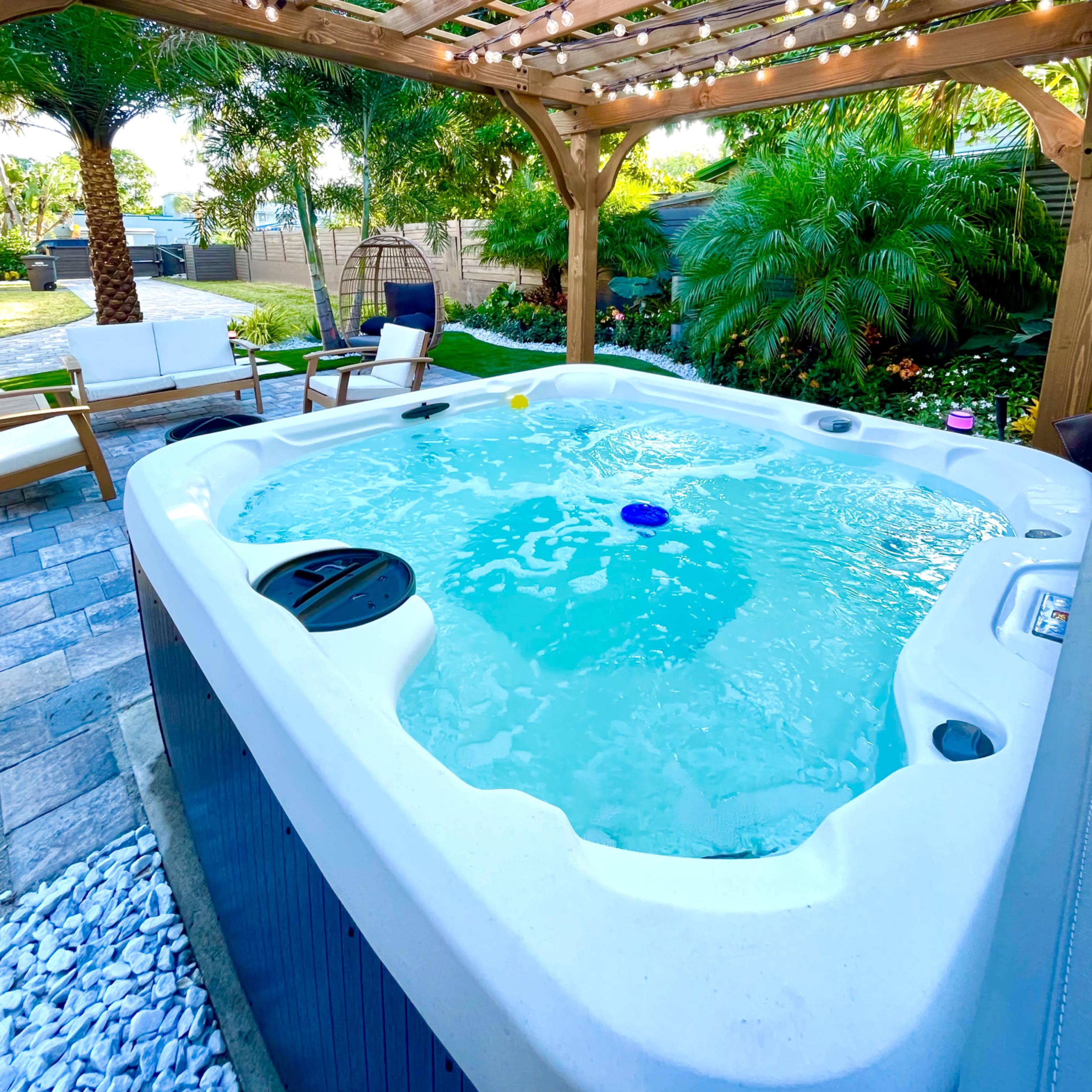 A hot tub is surrounded by a wooden pergola and tropical plants, with string lights overhead.