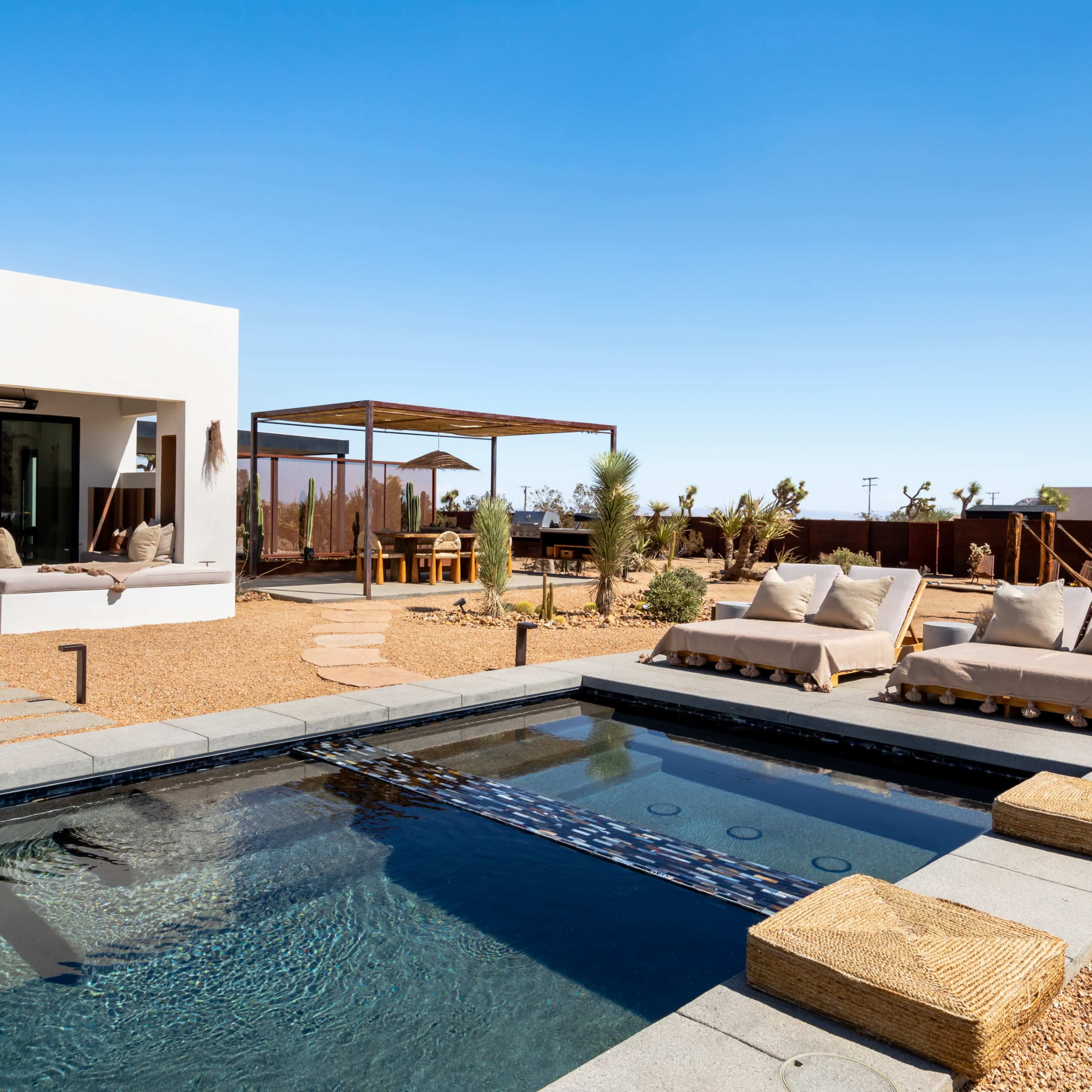 A modern residence features a swimming pool surrounded by lounge chairs and a shaded dining area in a desert landscape.