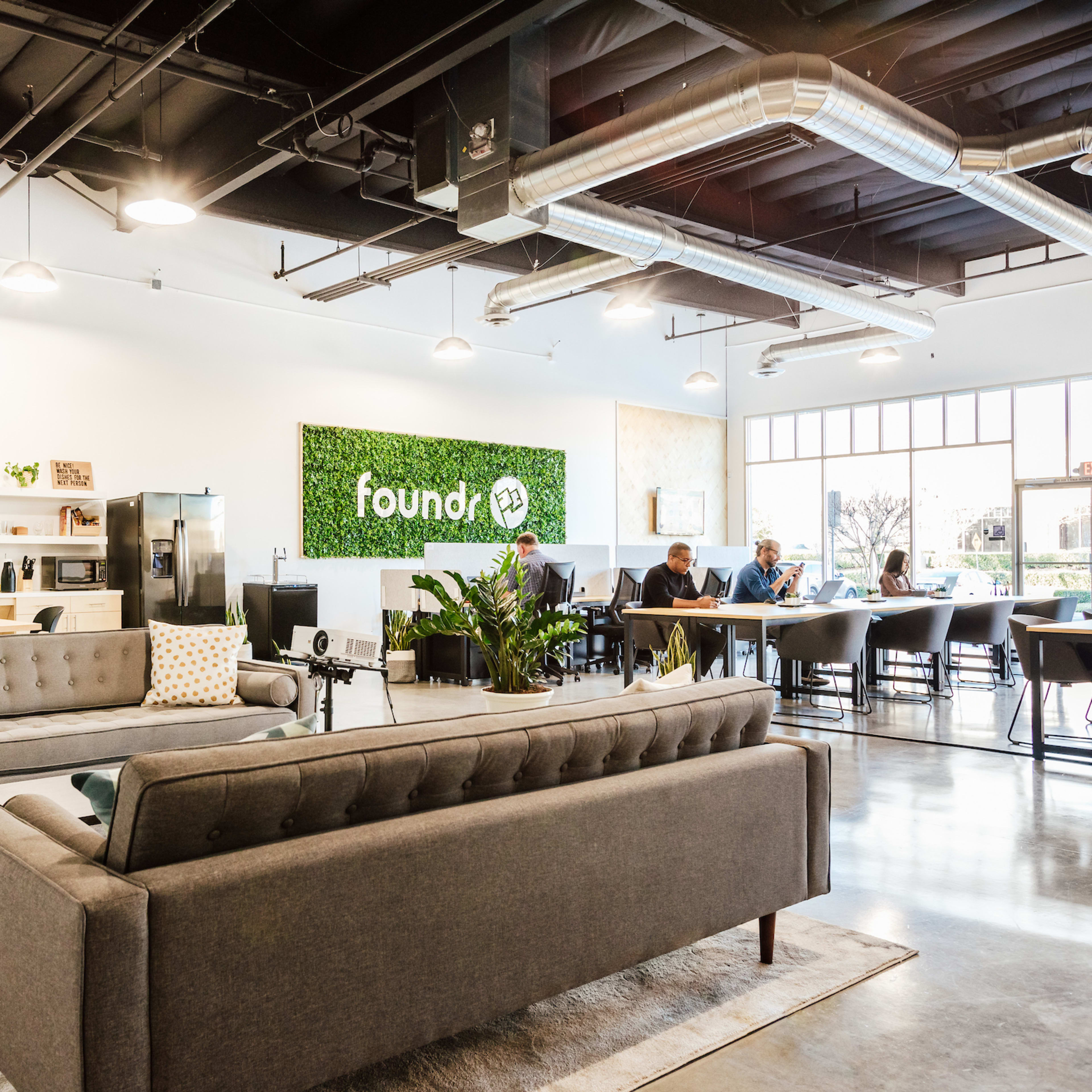 A modern lounge area with gray sofas, a green wall featuring the word "foundry," and several people working at tables in a bright, open space.
