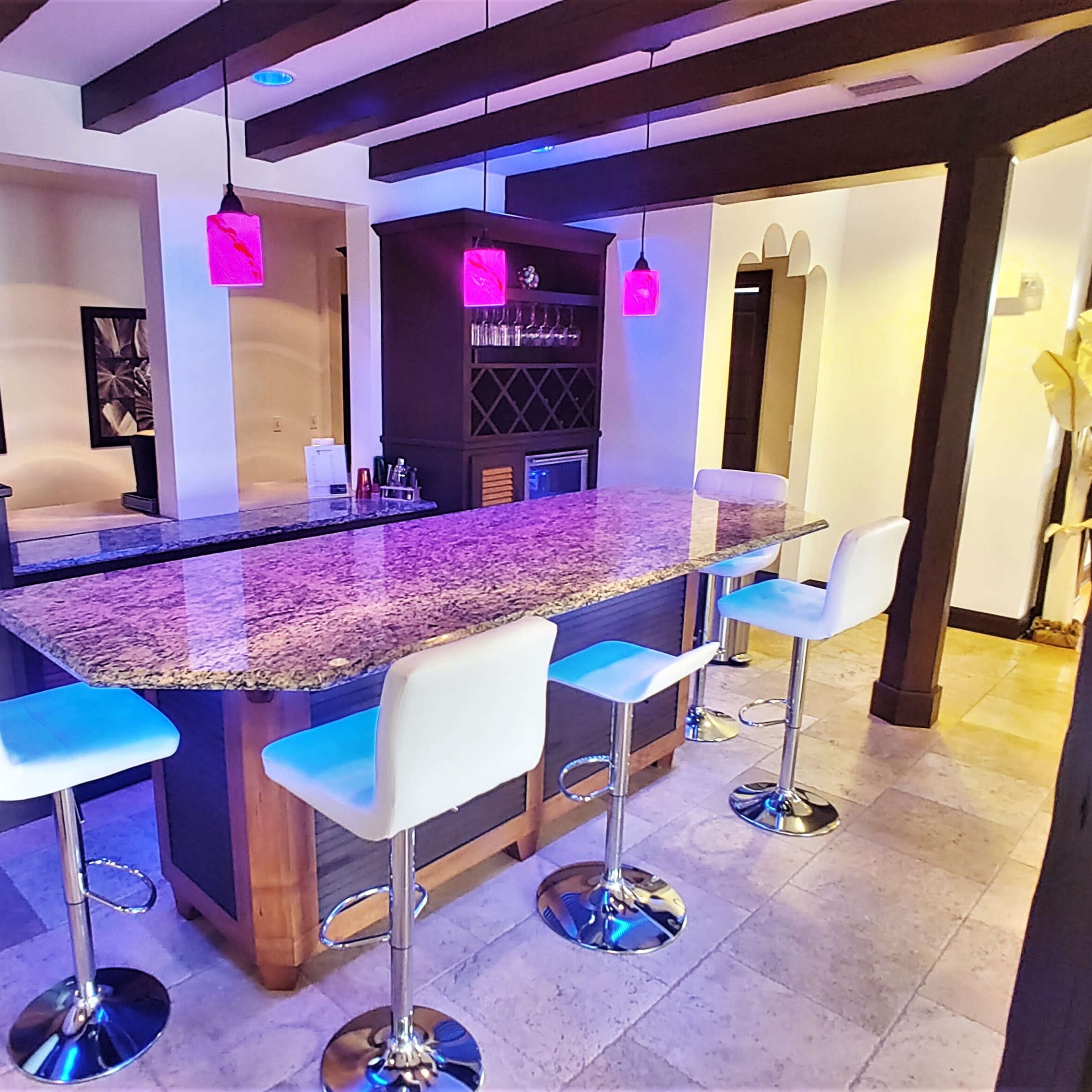 A modern kitchen bar area with a granite countertop, white high-backed stools, and colorful pendant lights hanging above.