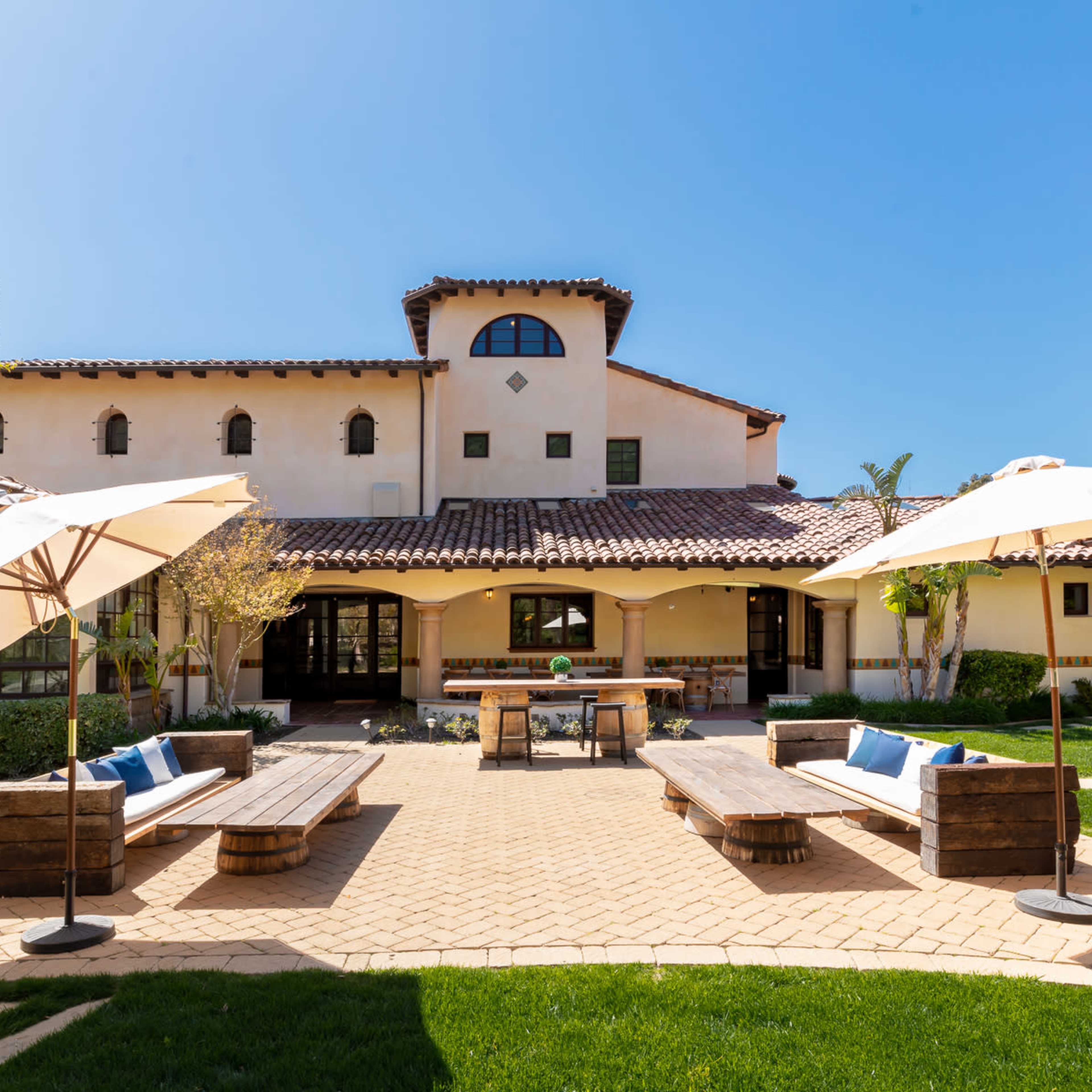 A spacious outdoor patio area with wooden benches and umbrellas, adjacent to a two-story house featuring a tiled roof and arched windows.