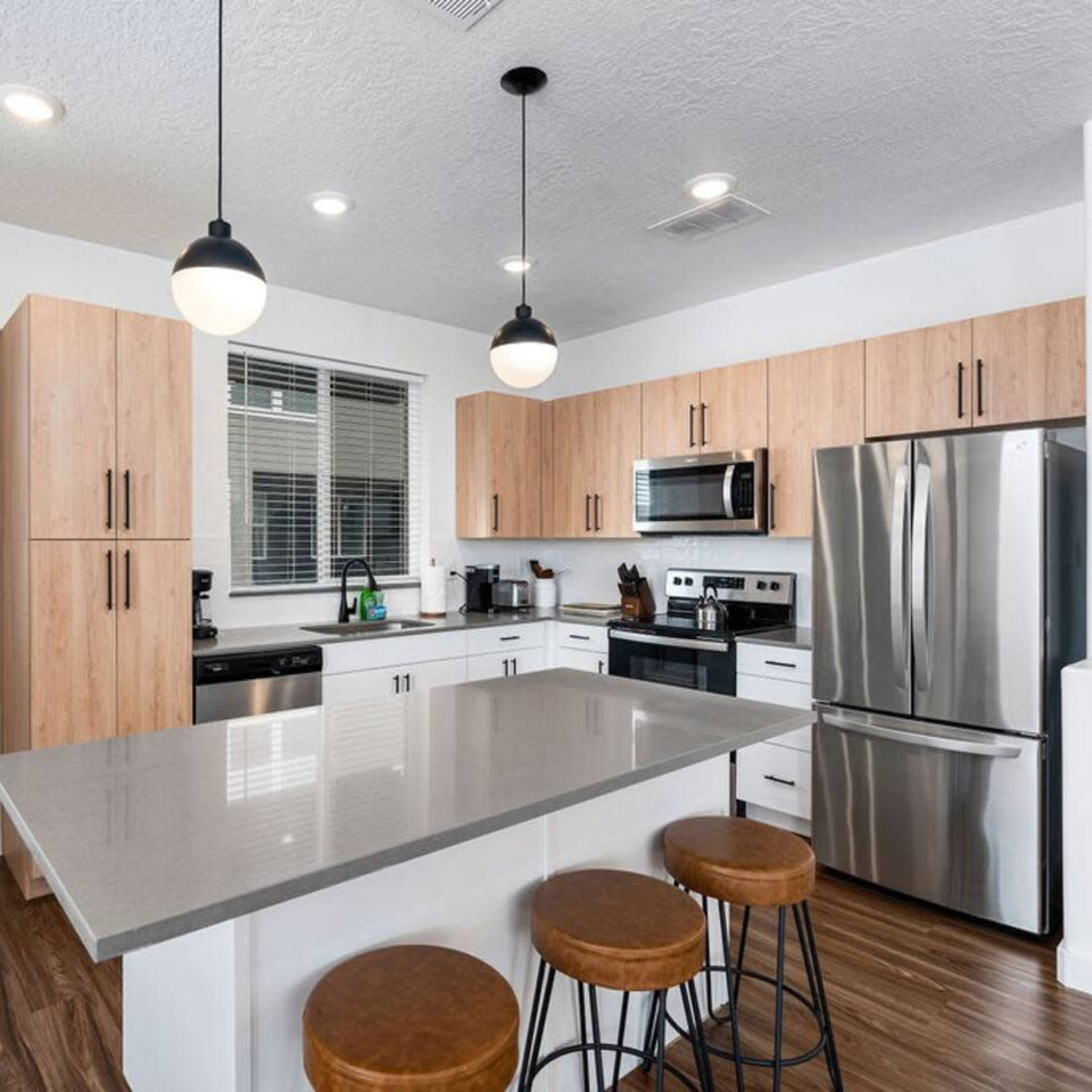 A modern kitchen with wooden cabinetry, stainless steel appliances, a gray countertop, and three bar stools at the island.