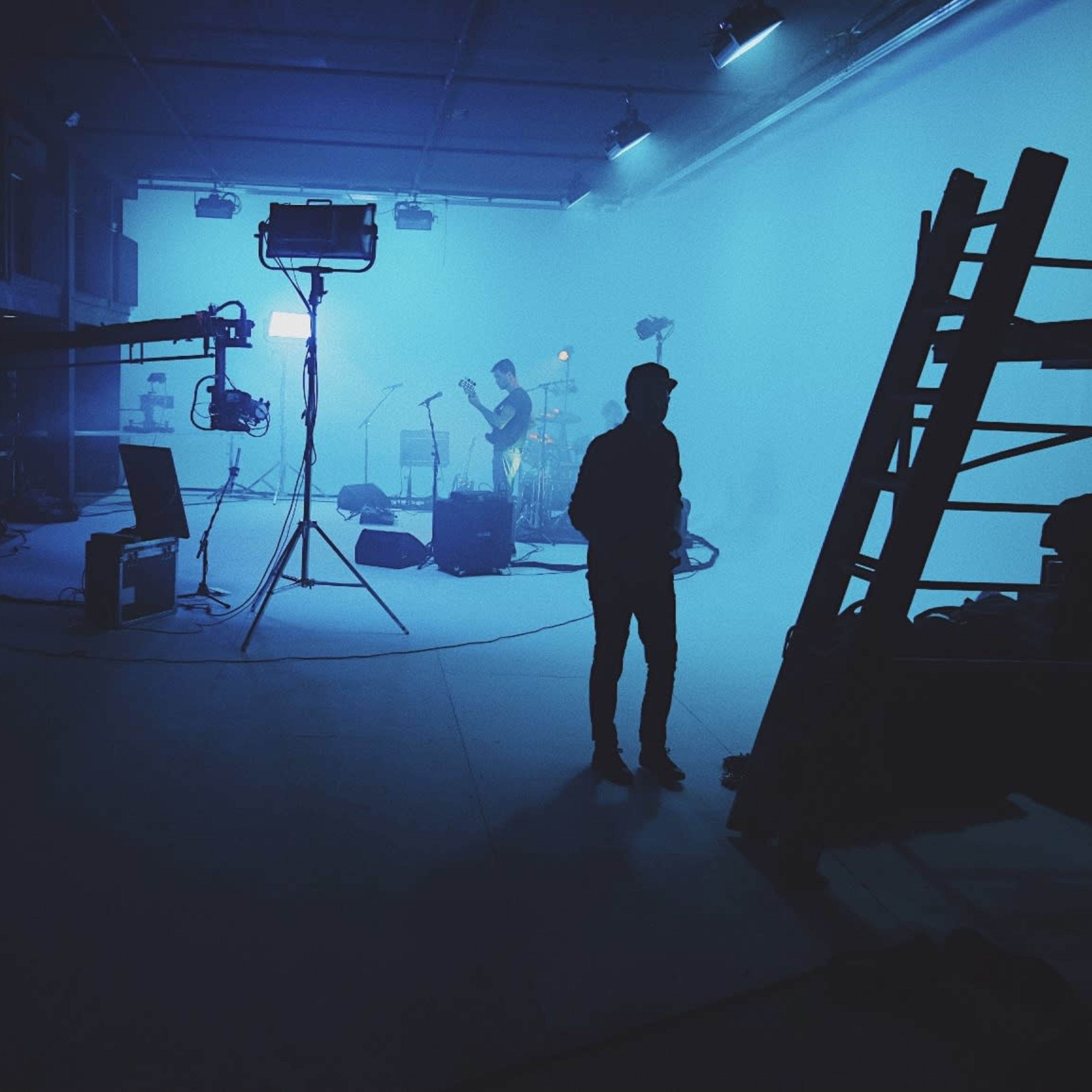 A silhouetted figure stands in a dimly lit studio while a band prepares to perform under blue lighting.