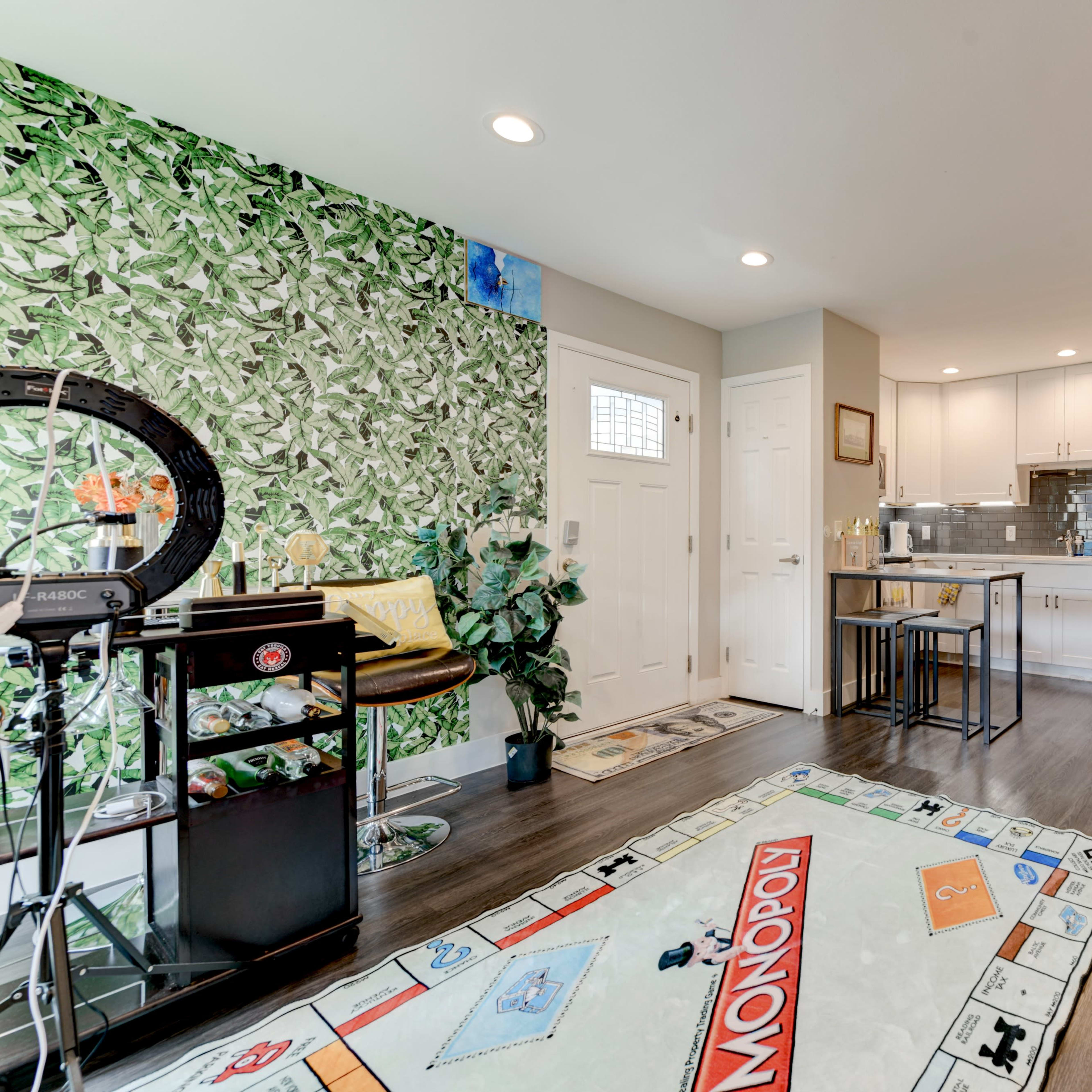 A modern living space featuring a kitchen area with a table, orange chairs, and a large Monopoly-themed rug on the floor.