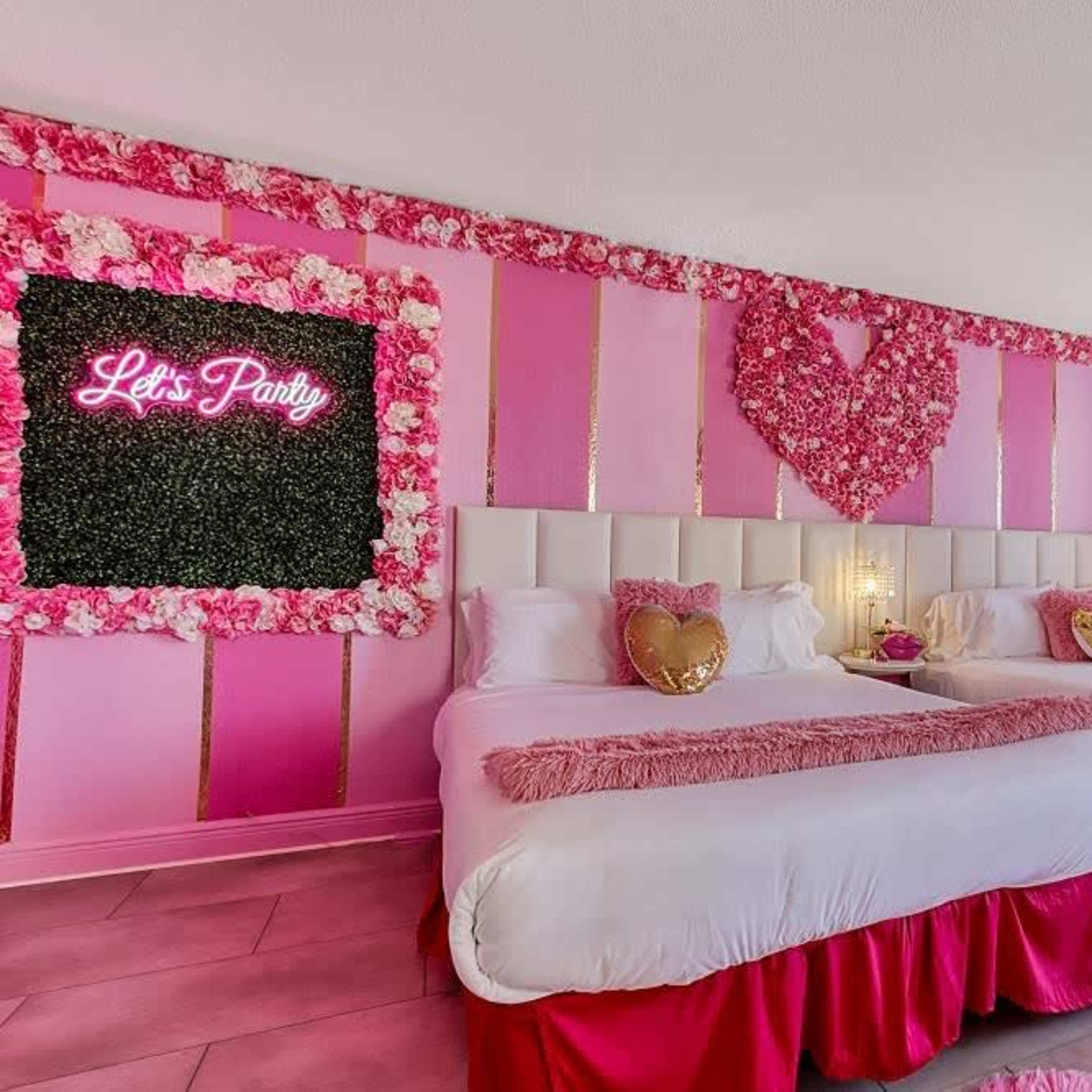 A brightly decorated hotel room with two beds, pink walls, floral accents, and a neon sign reading "Let's Party."