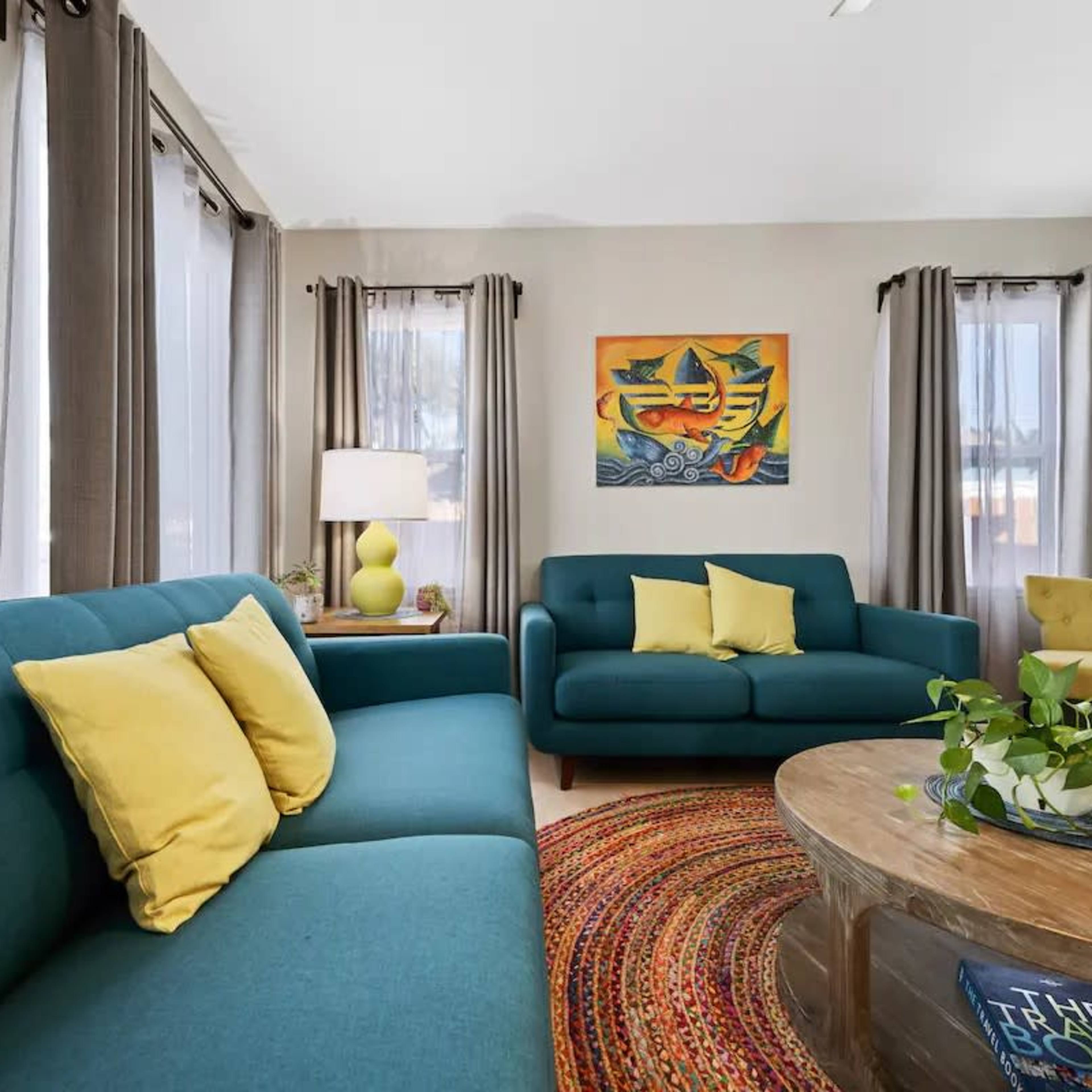 The living room features two teal sofas with yellow cushions, a round wooden coffee table, and a colorful painting on the wall, accented by natural light through the curtains.